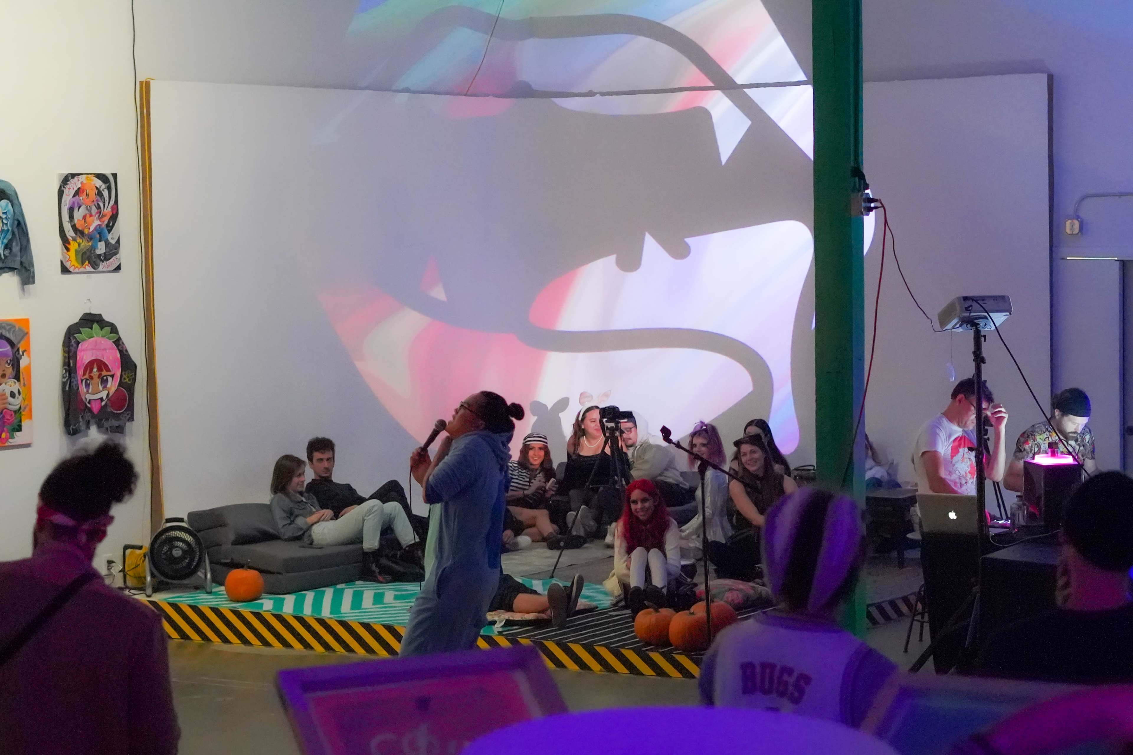 The scene shows a vibrant art event with people sitting on a patterned floor, a performer in a onesie singing, and colorful projections on a white wall.