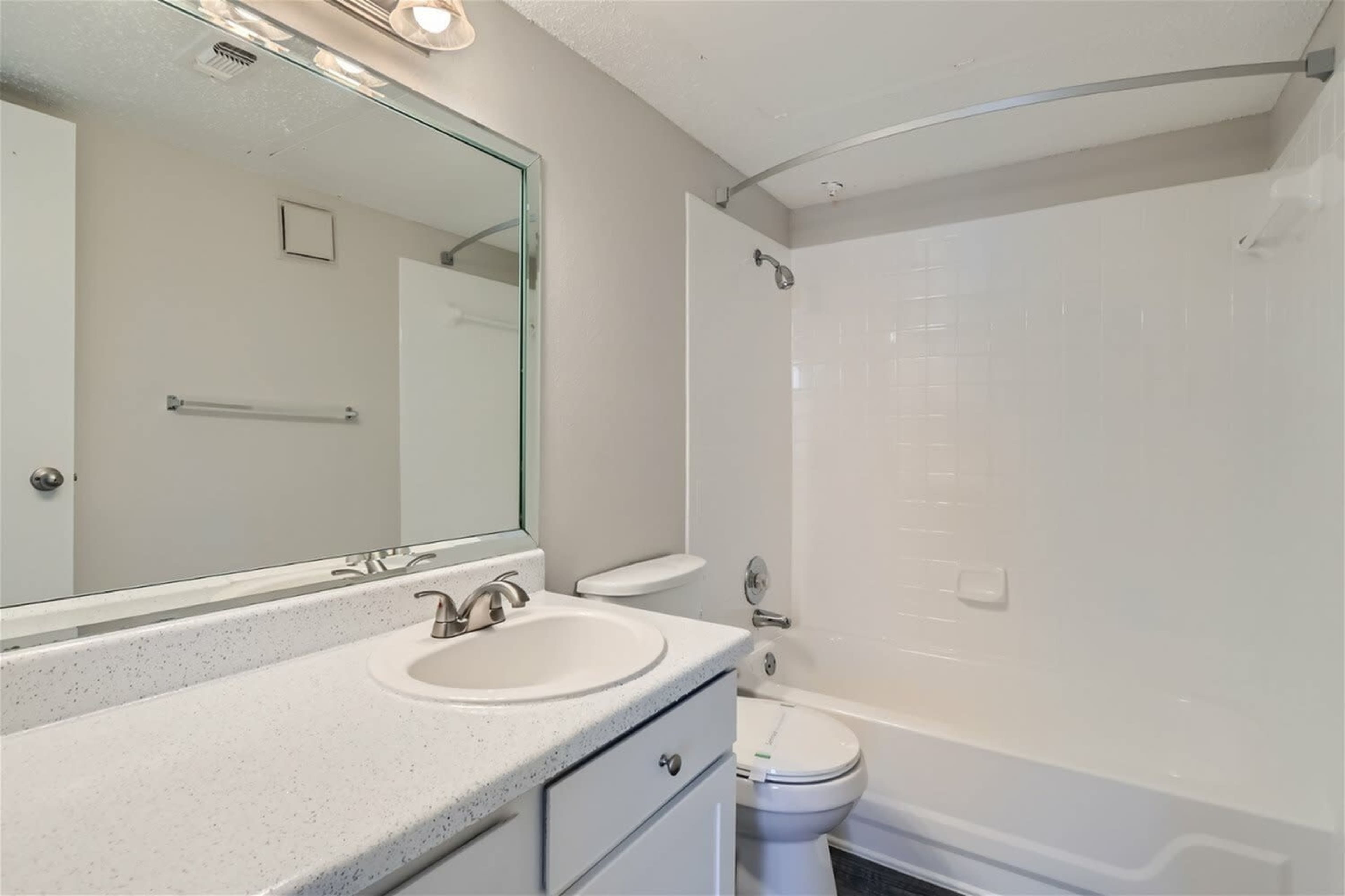 The image shows a clean bathroom featuring a bathtub with a shower, a white sink with a countertop, and a large mirror above the sink.