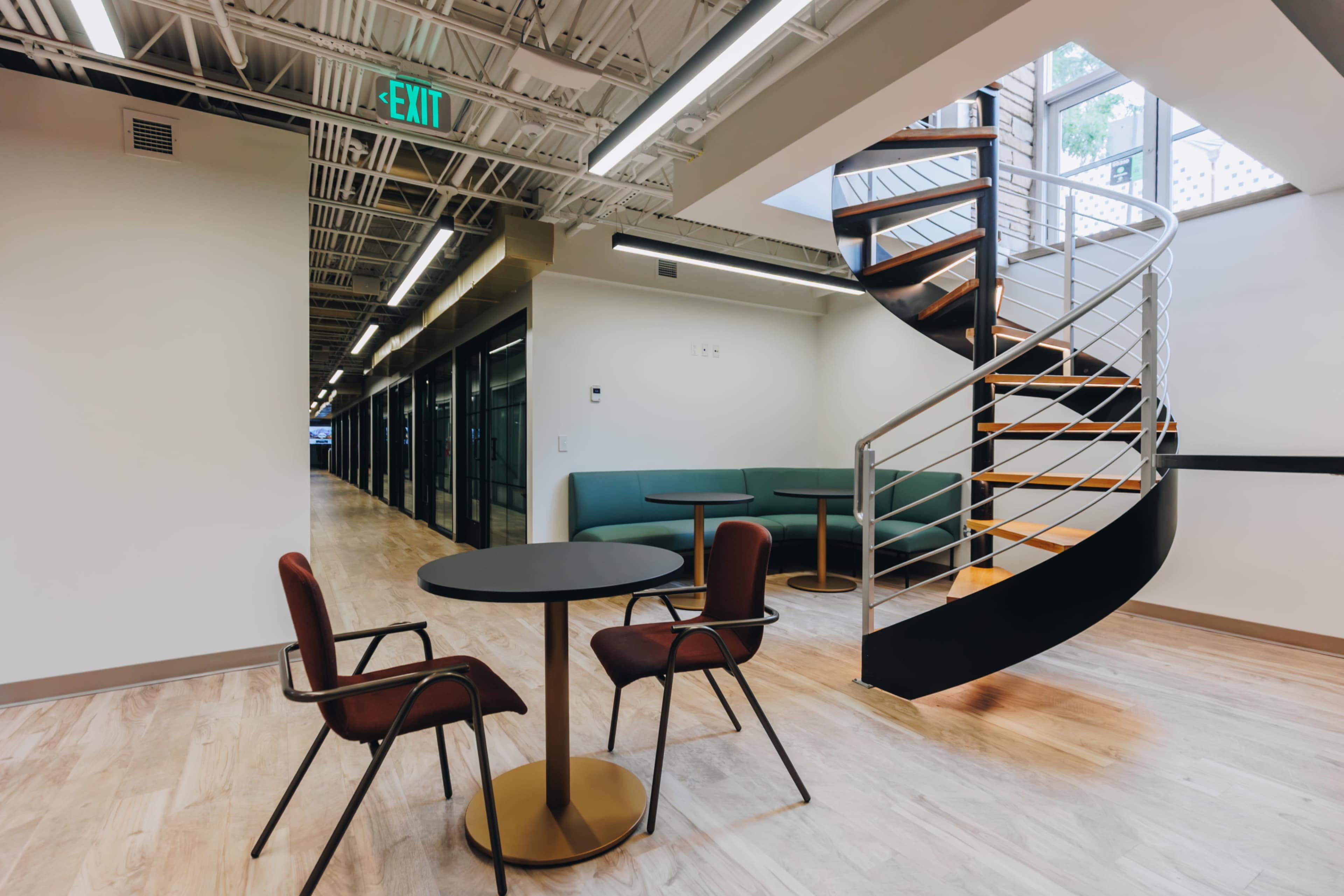 The image shows a modern office space featuring a spiral staircase, circular table with two chairs, and glass-walled meeting rooms.