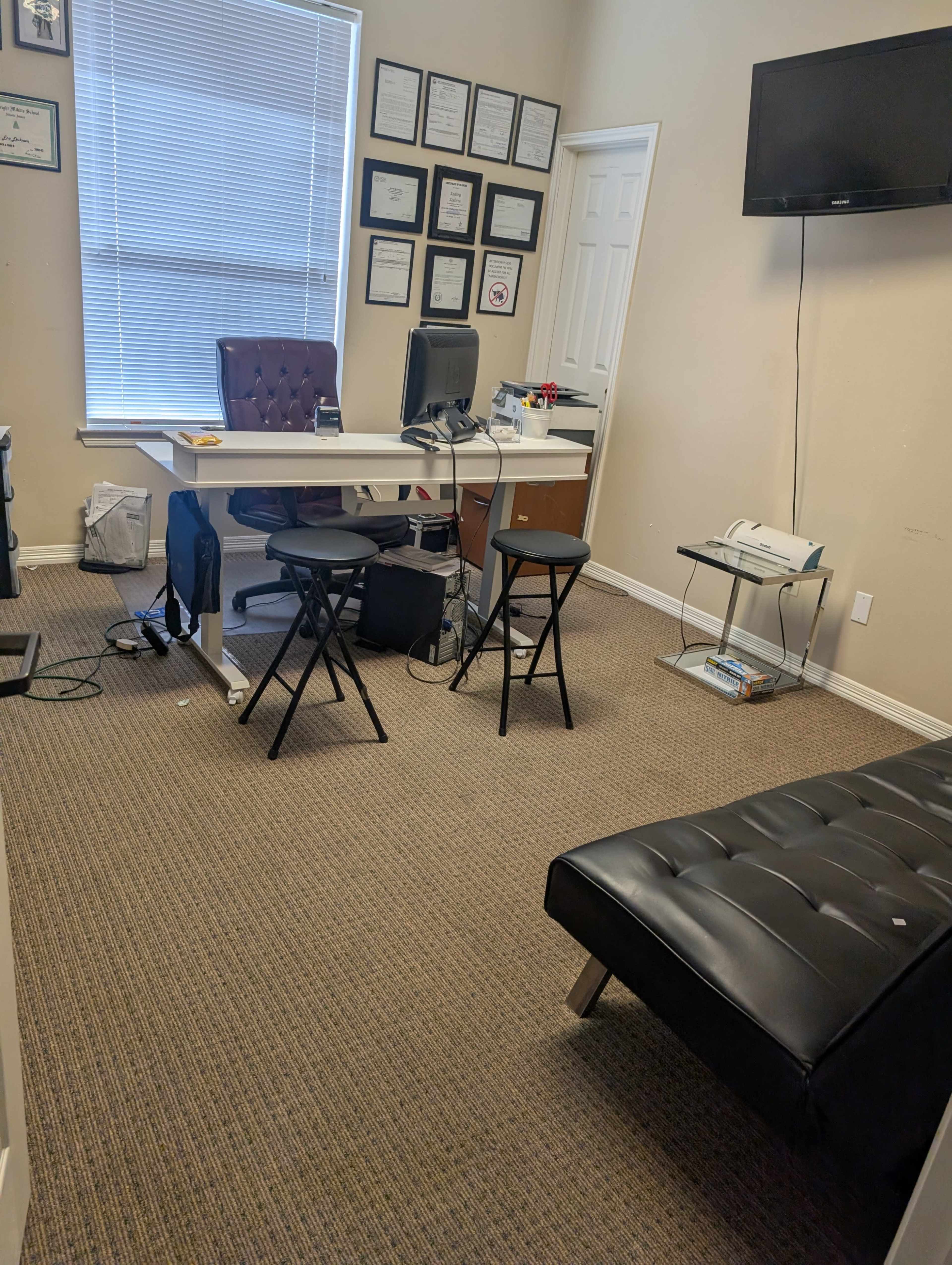 The image shows a small office space with a desk, chair, and a television mounted on the wall, along with a futon and various framed documents.