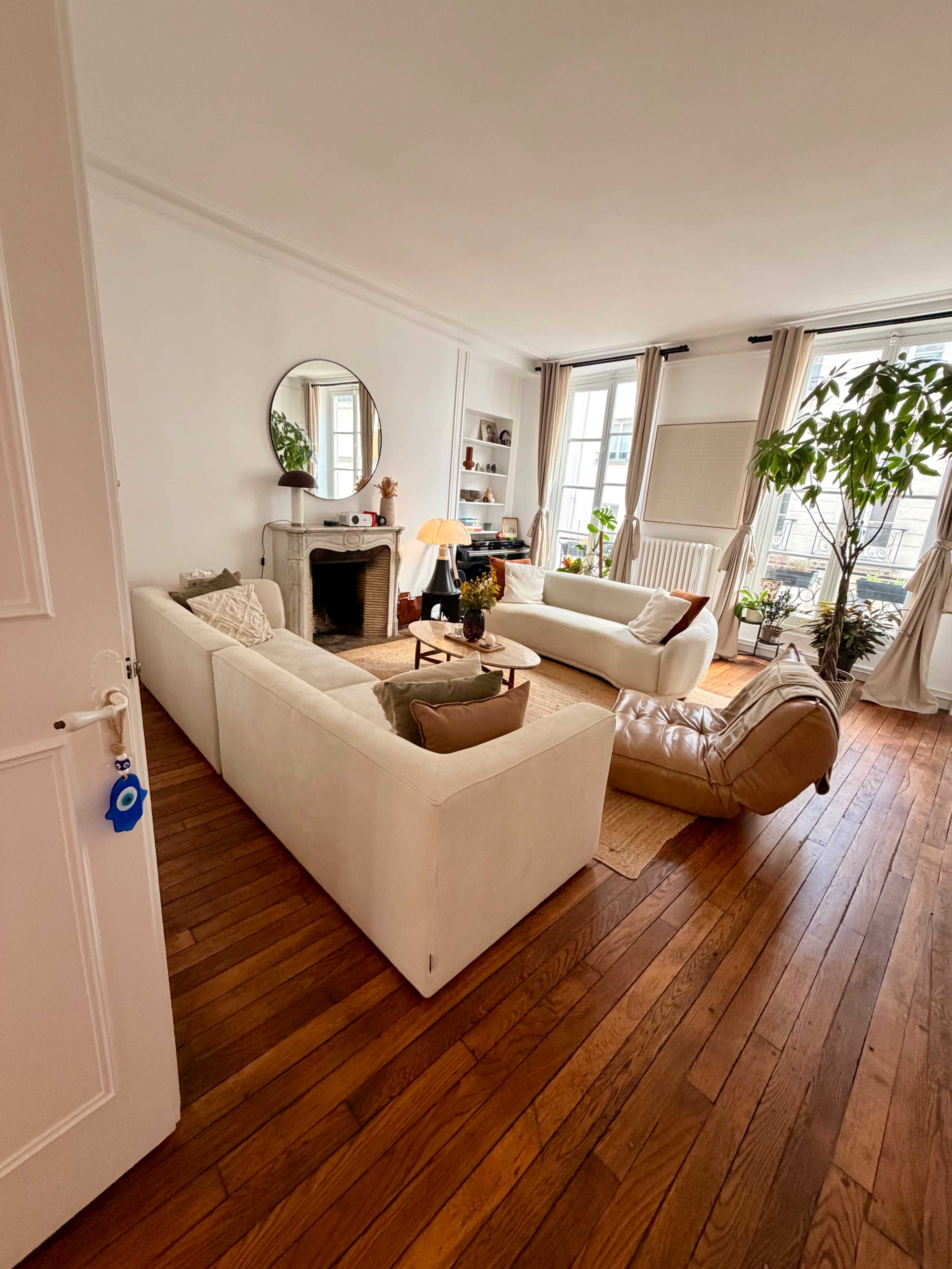 The image shows a bright living room featuring two white sofas, a round mirror, a small fireplace, and indoor plants arranged around a wooden floor.