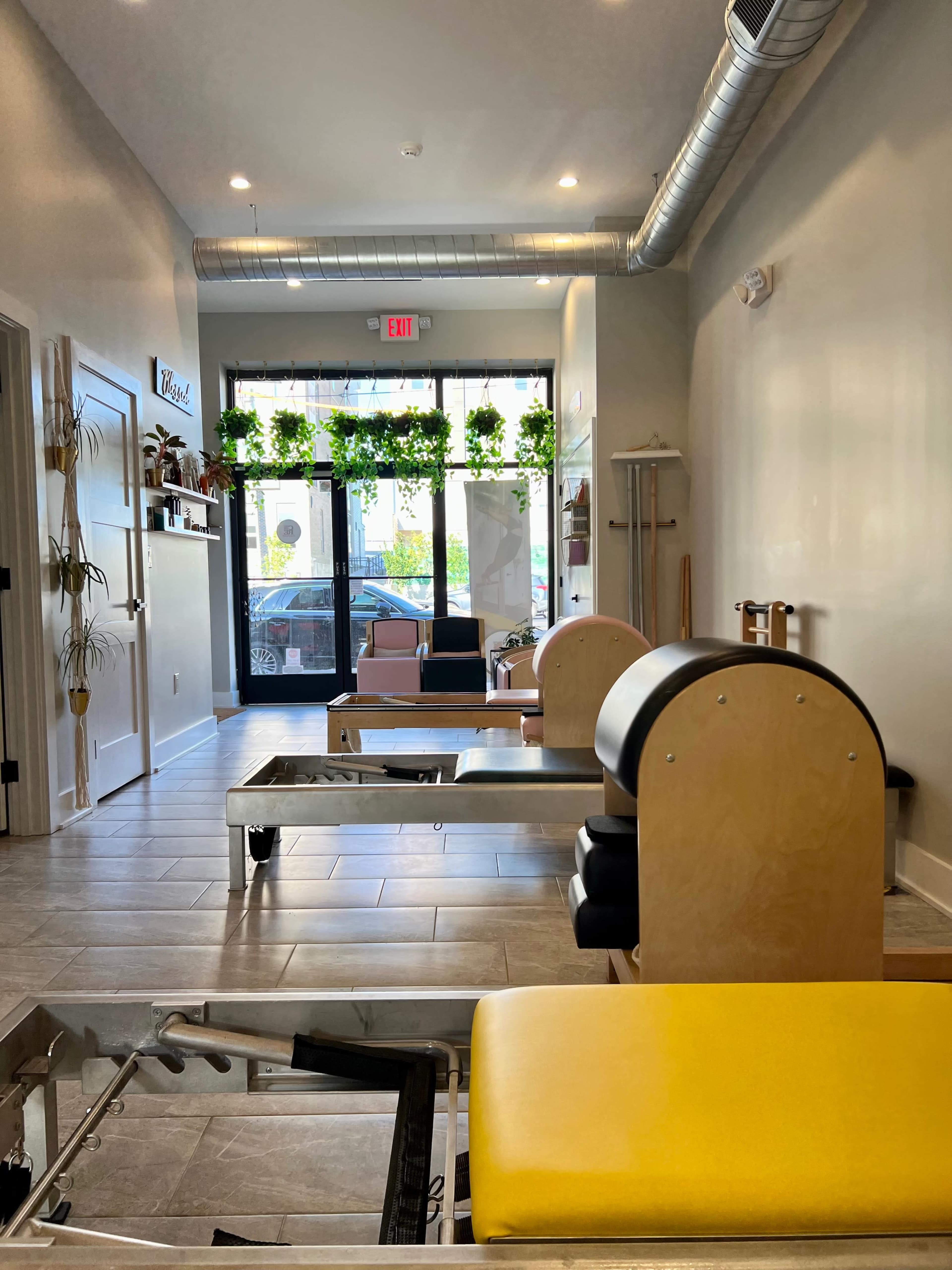 The interior of a studio featuring several Pilates reformers, with large windows displaying plants and natural light.