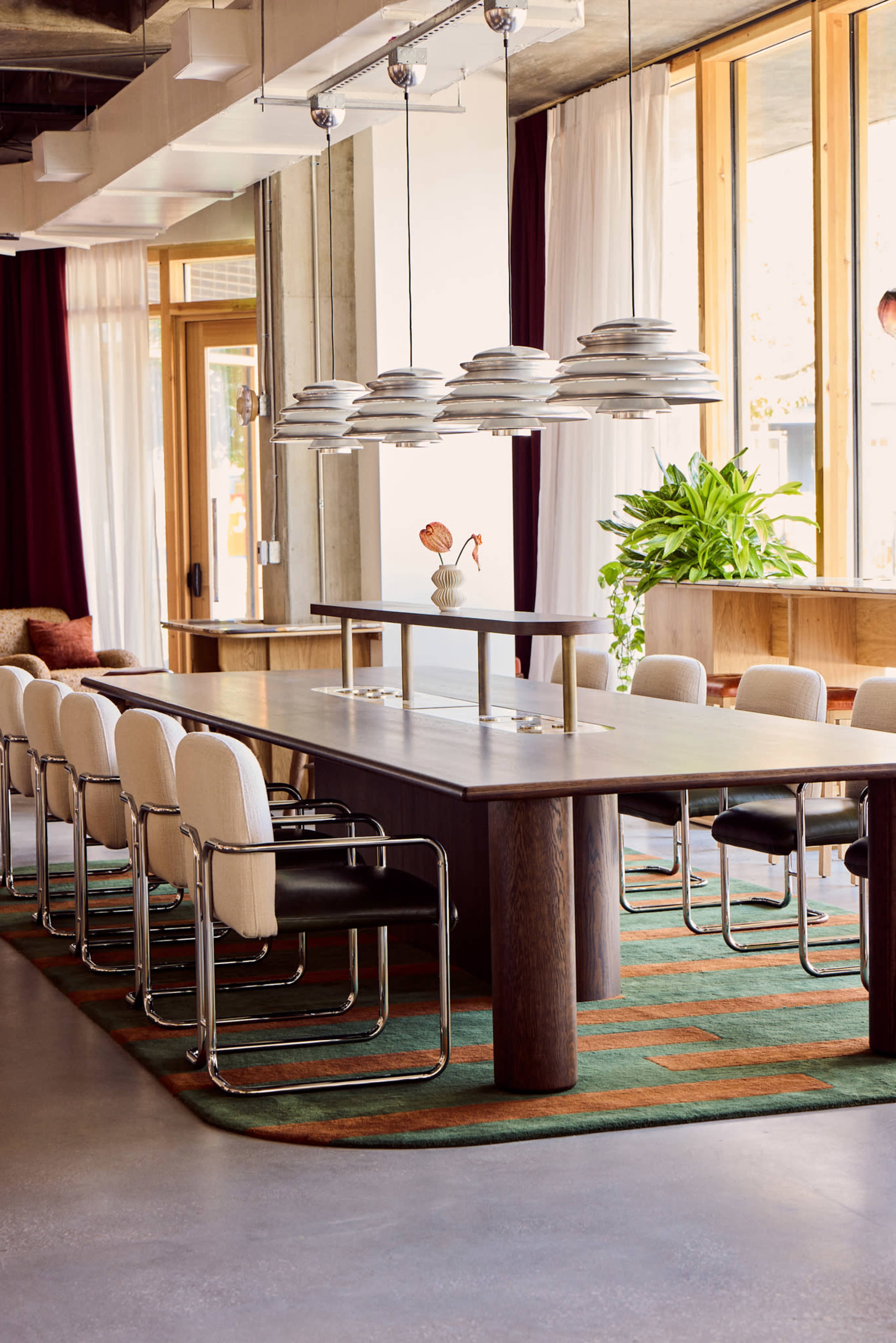 The image shows a modern dining area featuring a long wooden table surrounded by white and black chairs, with pendant lights hanging above and large windows letting in natural light.