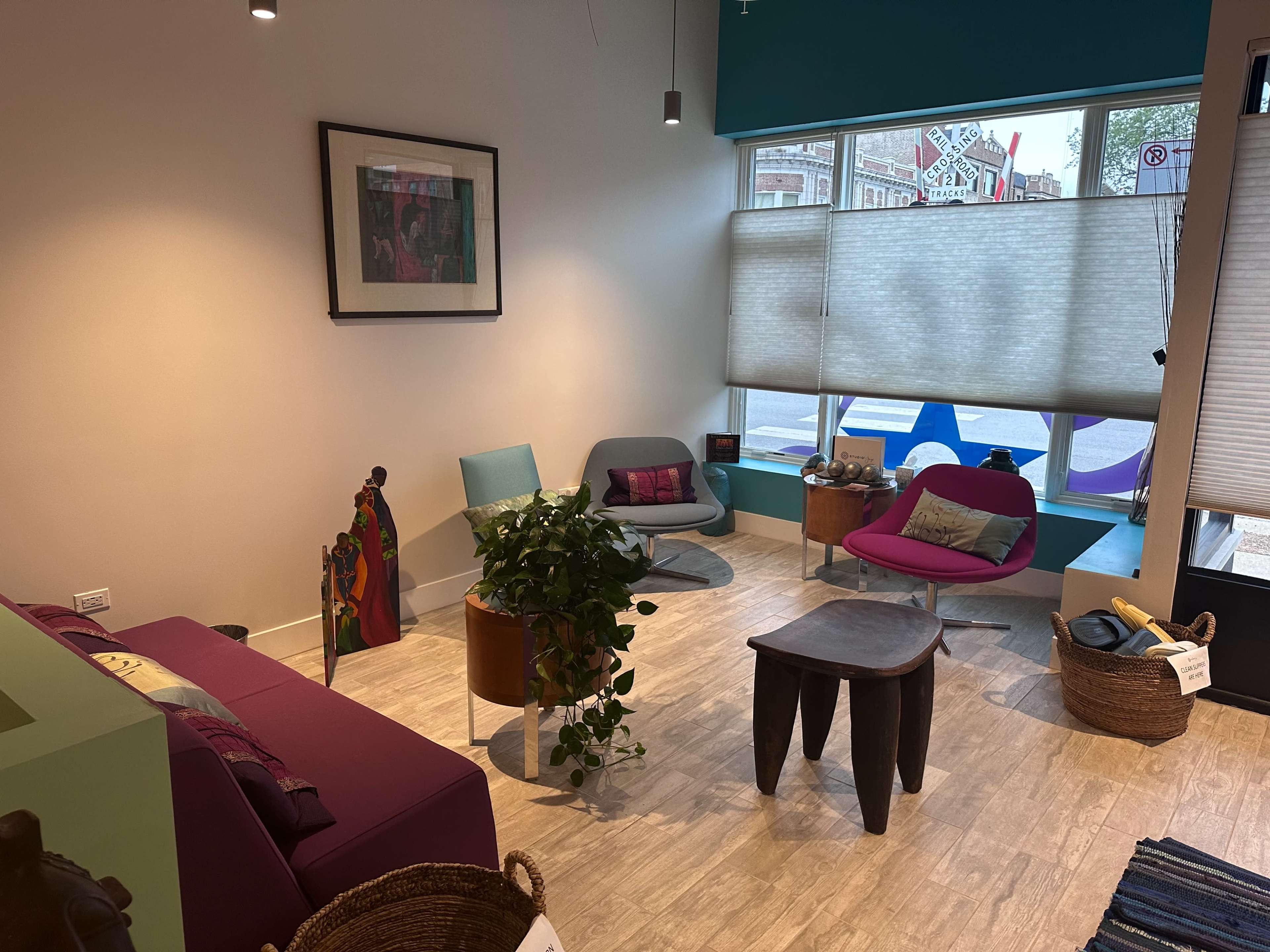 The image shows a modern, minimalist seating area with colorful chairs, a coffee table, and a potted plant beside large windows with blinds.