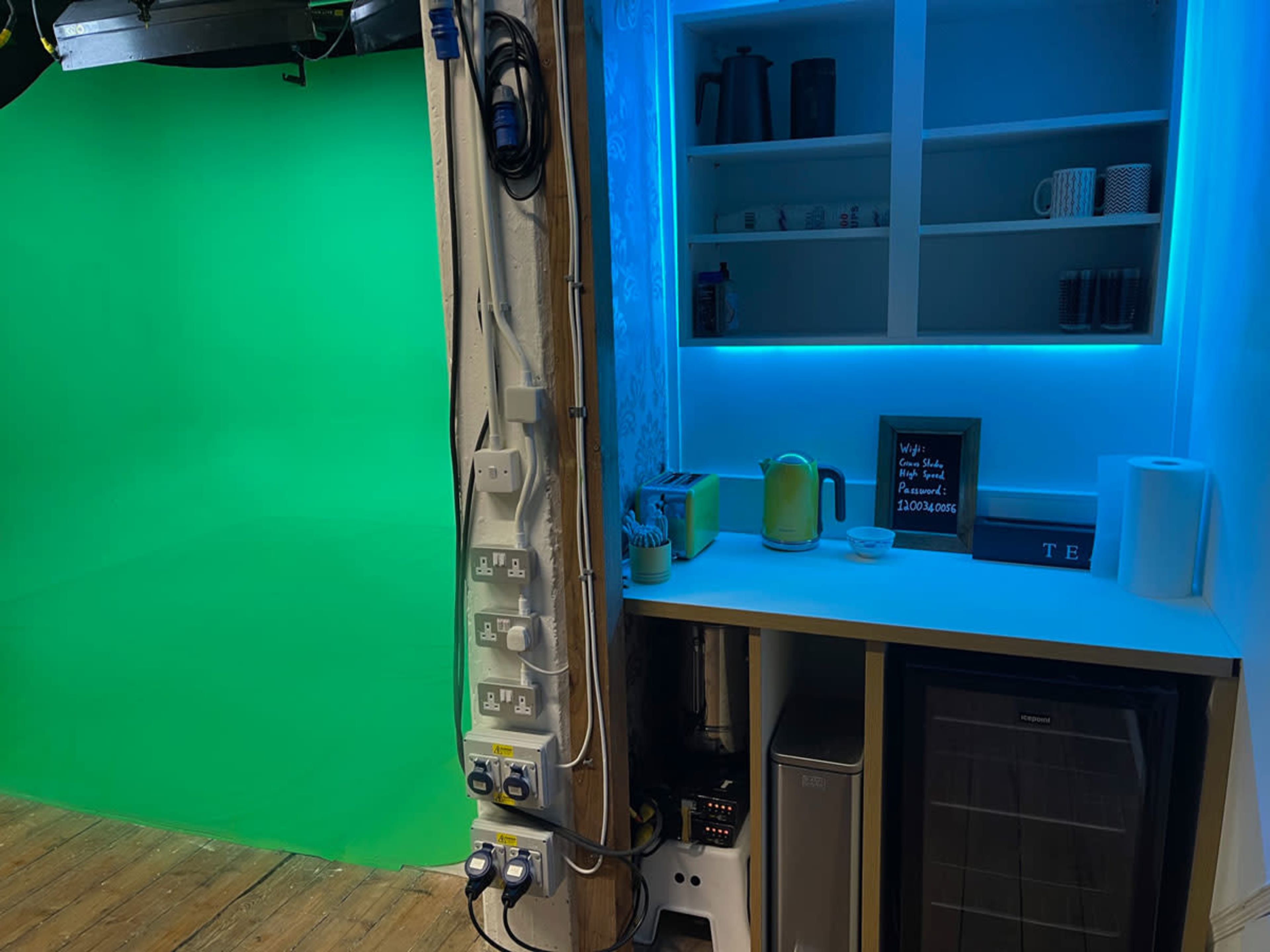 A studio setup with a green screen on one side and a small kitchenette featuring a refrigerator, sink, and various kitchen items on the other side.
