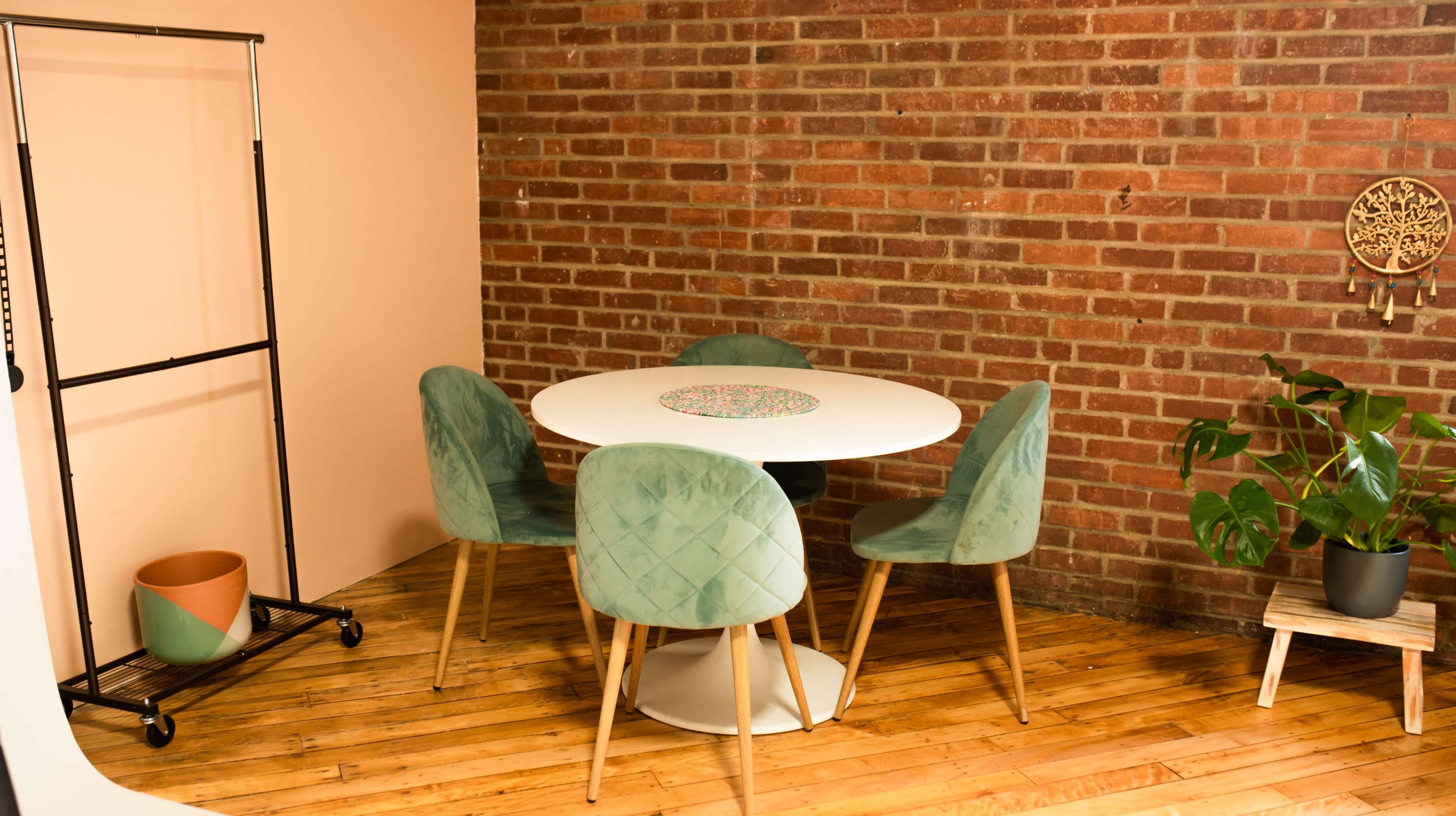 A round white table surrounded by four green chairs sits on a wooden floor against a brick wall, with a potted plant in the corner.