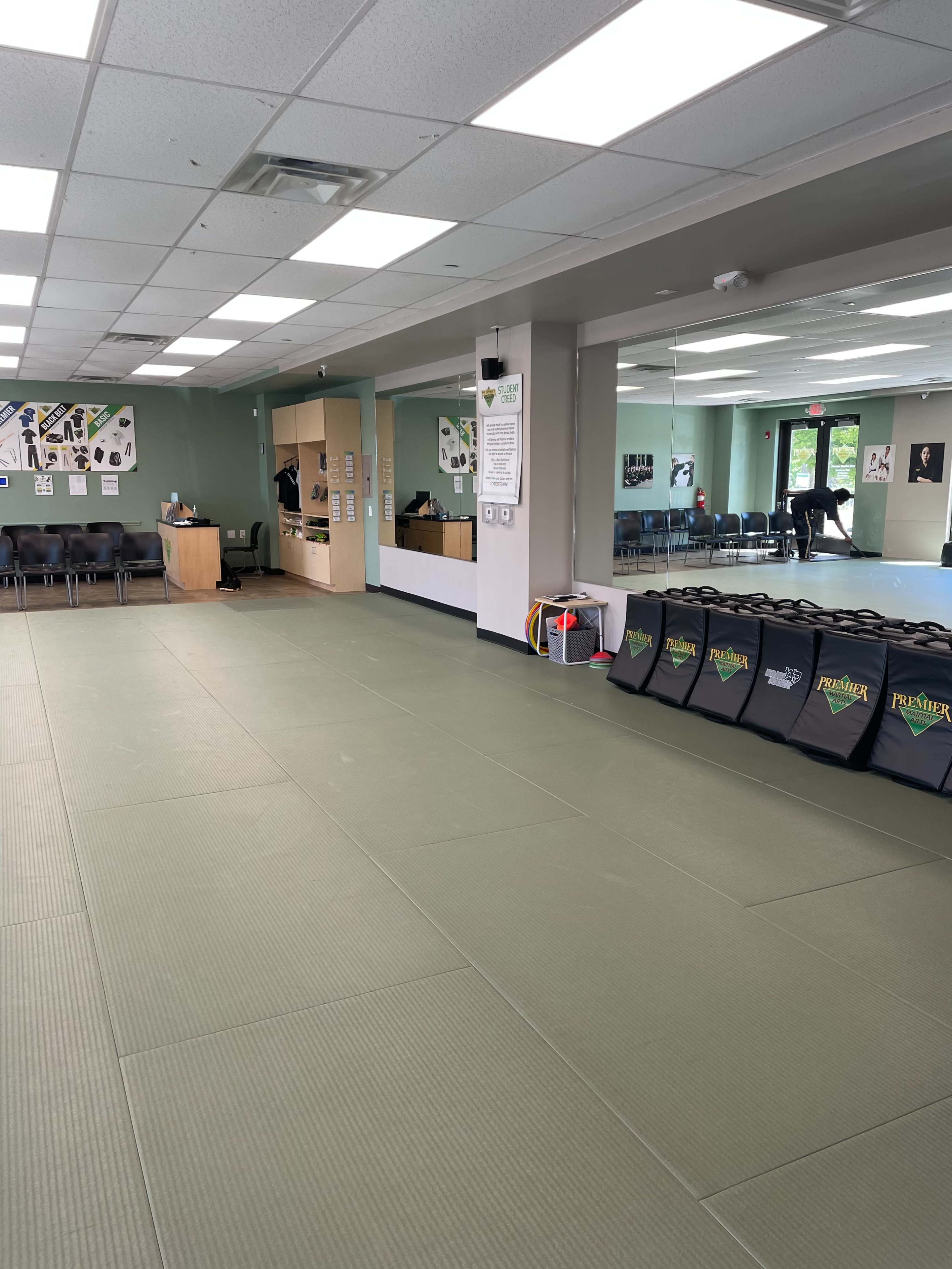 The image shows a spacious martial arts studio with padded flooring, a large mirror, and neatly arranged training mats along one wall.