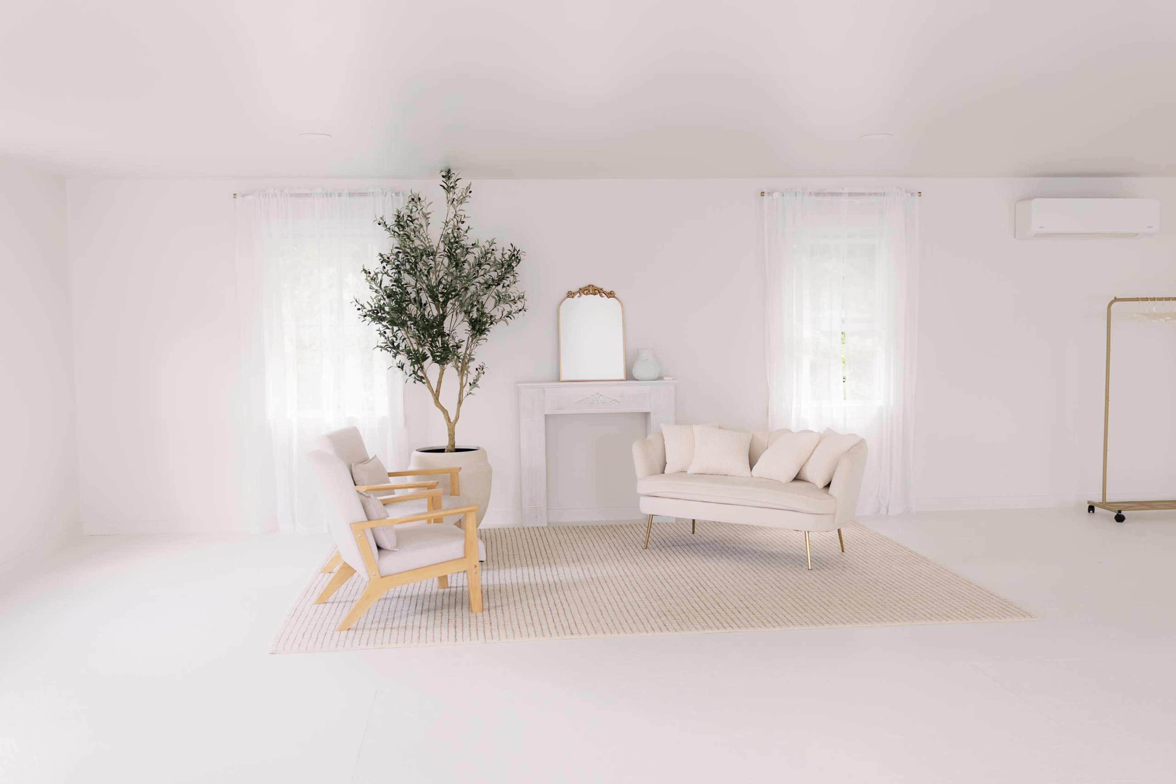 A modern, minimalist living space features two light wooden armchairs and a cream-colored sofa arranged on a neutral rug, with a decorative tree, a mirror, and a fireplace in the background.