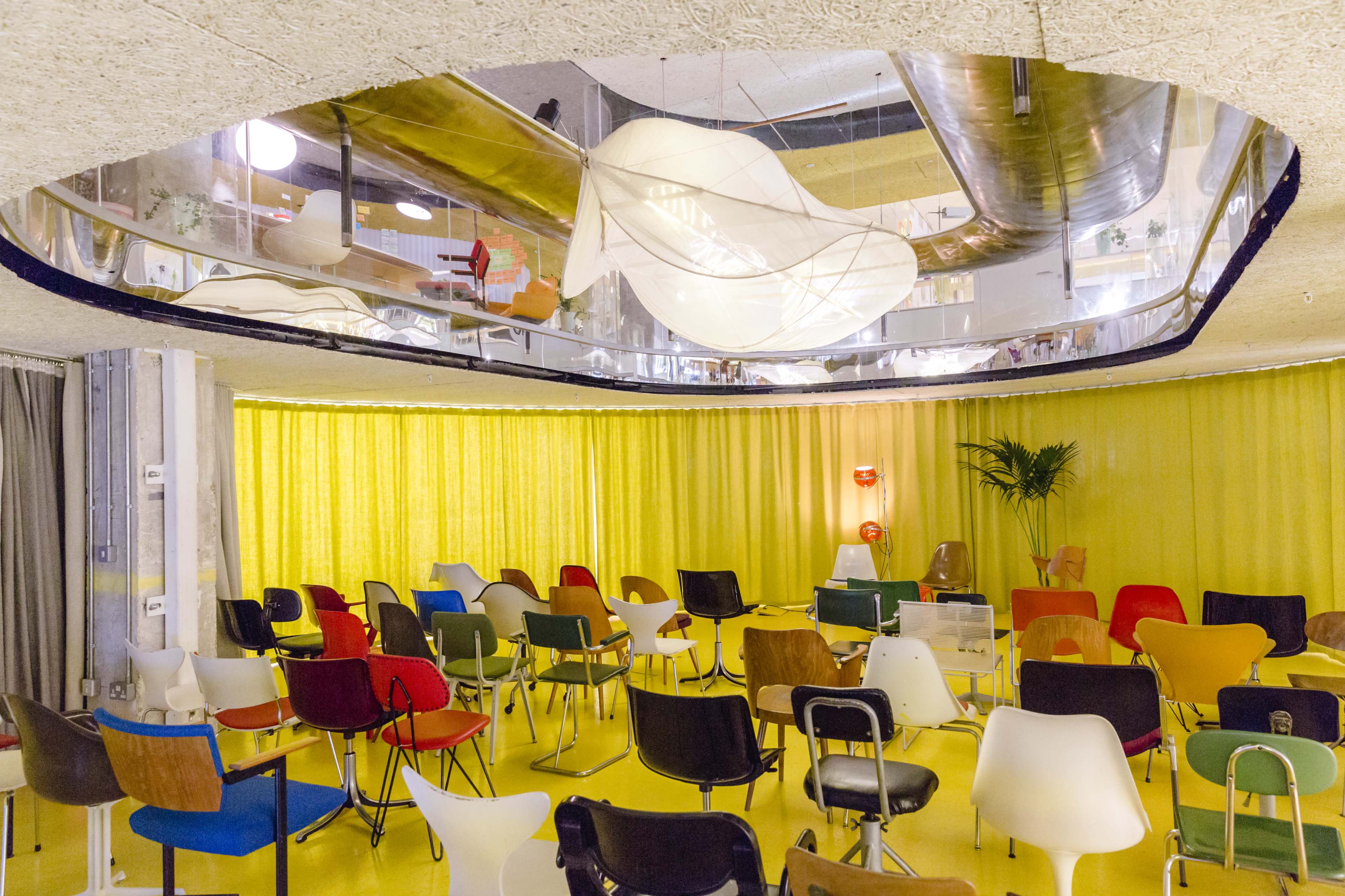 A brightly lit room features rows of colorful chairs arranged in a circular formation beneath a mirrored ceiling.