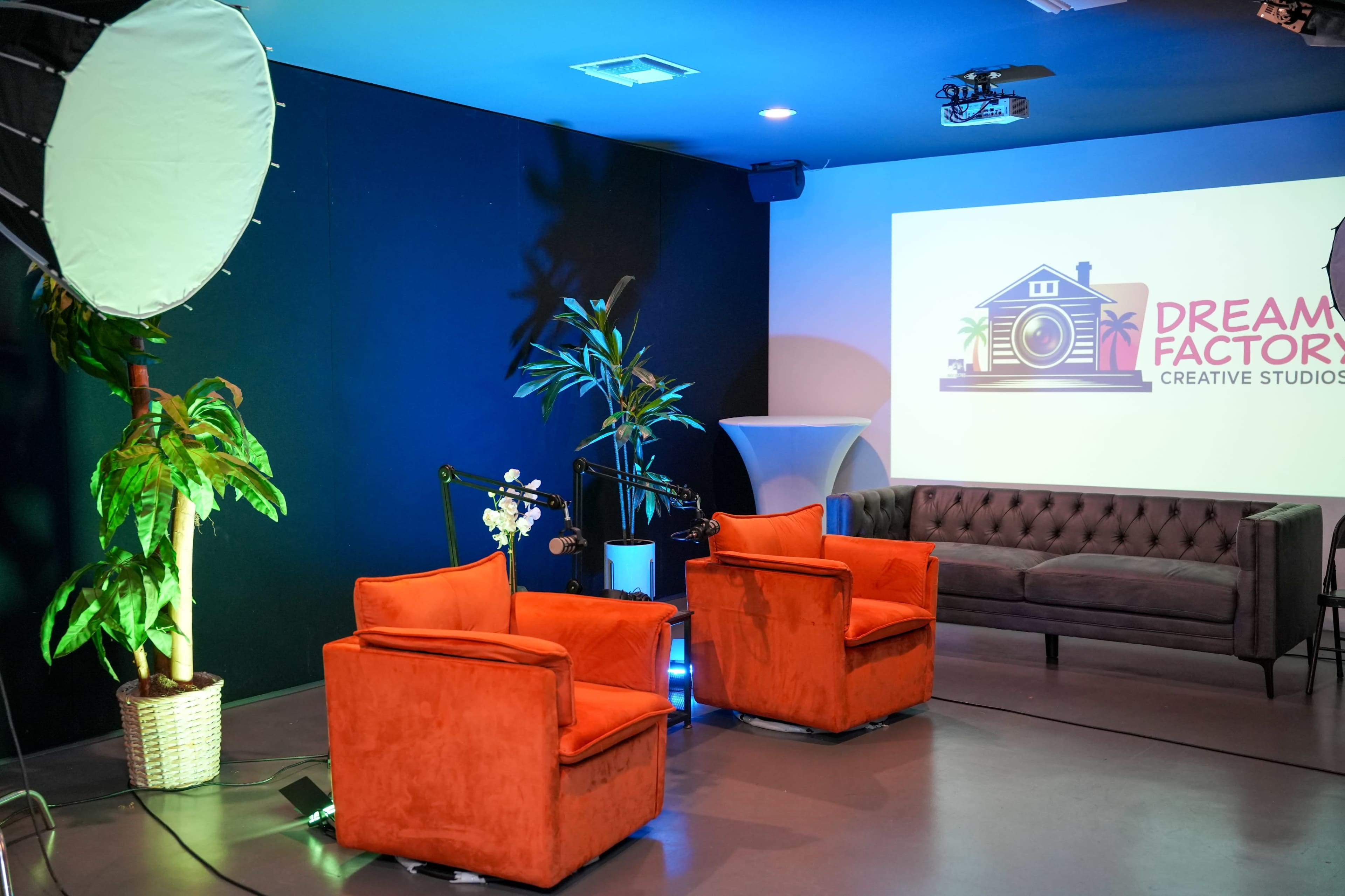 The image shows a modern studio set with two orange armchairs, a backdrop featuring a large logo for "Dream Factory Creative Studios," and a plant in the corner.