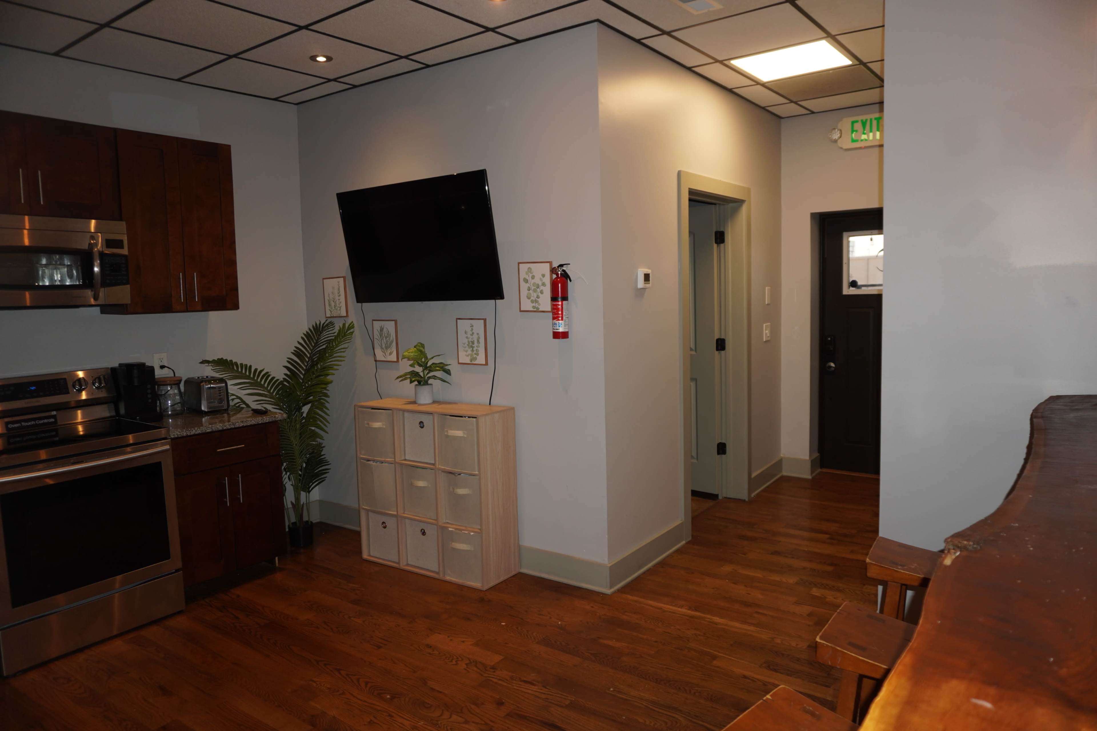 The image shows a contemporary interior space featuring a small kitchen area with stainless steel appliances, a wooden shelf, and a television mounted on the wall, leading into a hallway with a door and a potted plant.