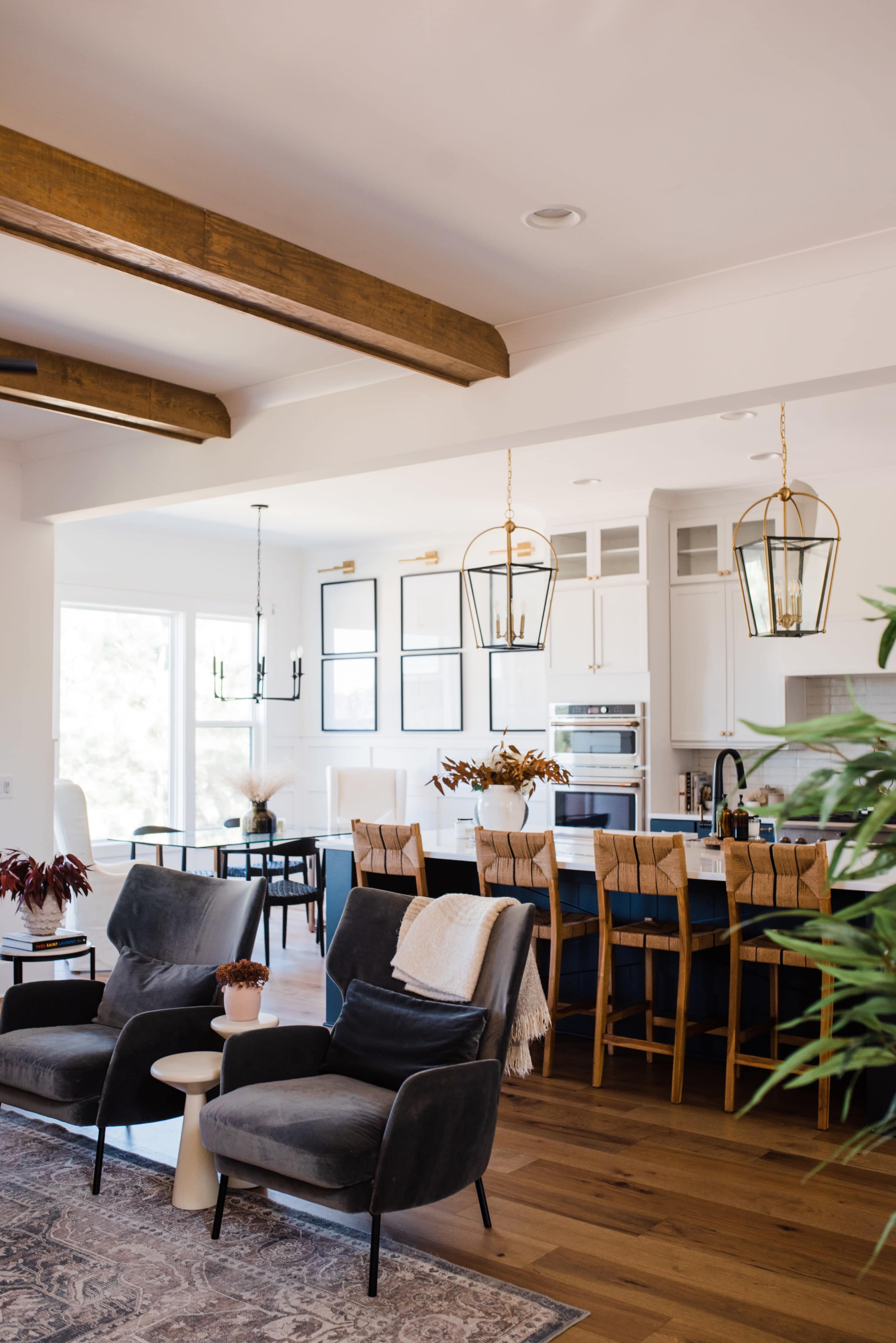 A modern kitchen and living area feature a large island with seating, hanging light fixtures, and dark chairs arranged around a table.