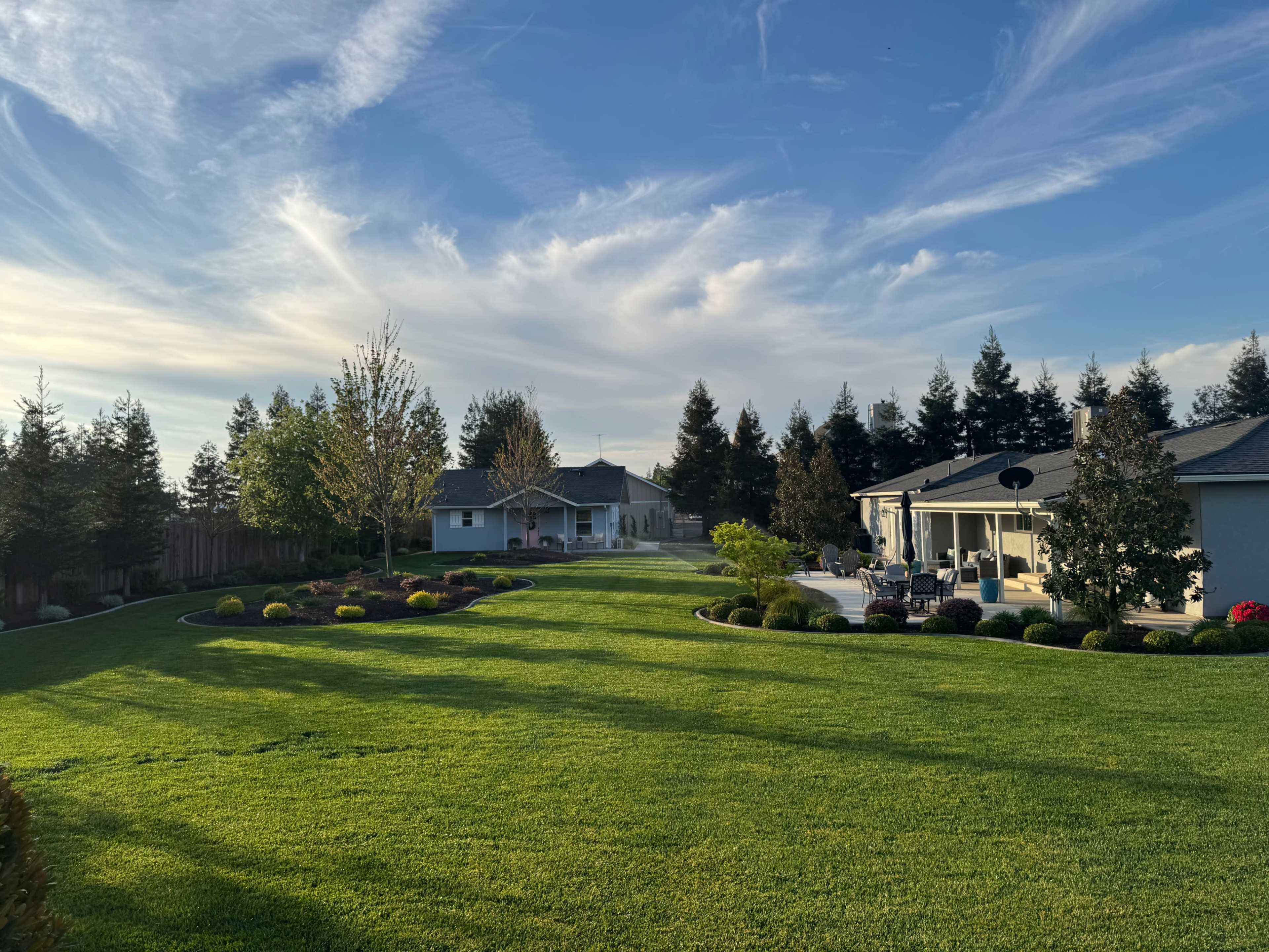 The image shows a well-maintained lawn with landscaped gardens, a house in the background, and trees lining the perimeter under a blue sky with wispy clouds.