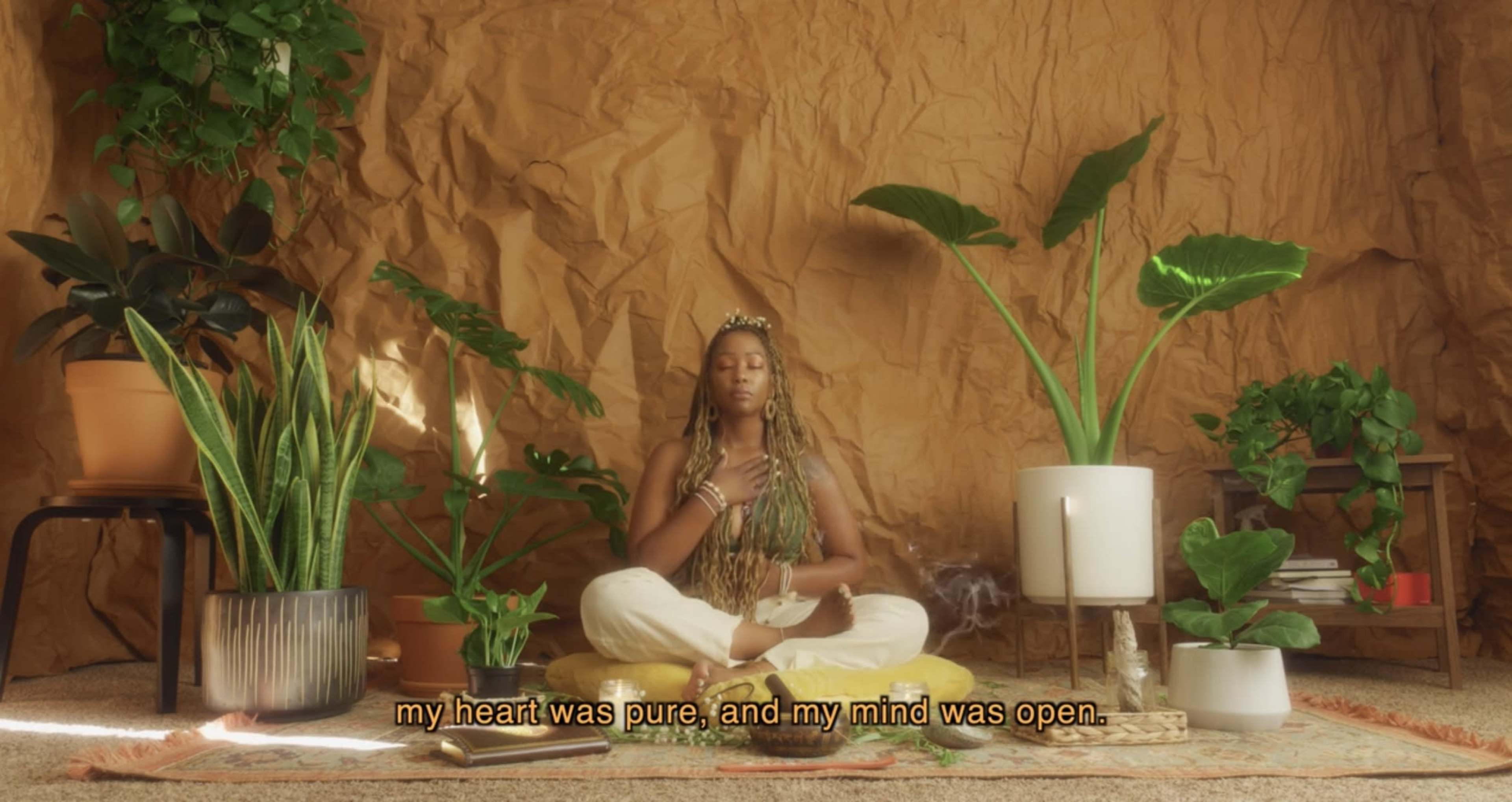 A person sits cross-legged on the floor surrounded by various potted plants against a textured, earthy backdrop.