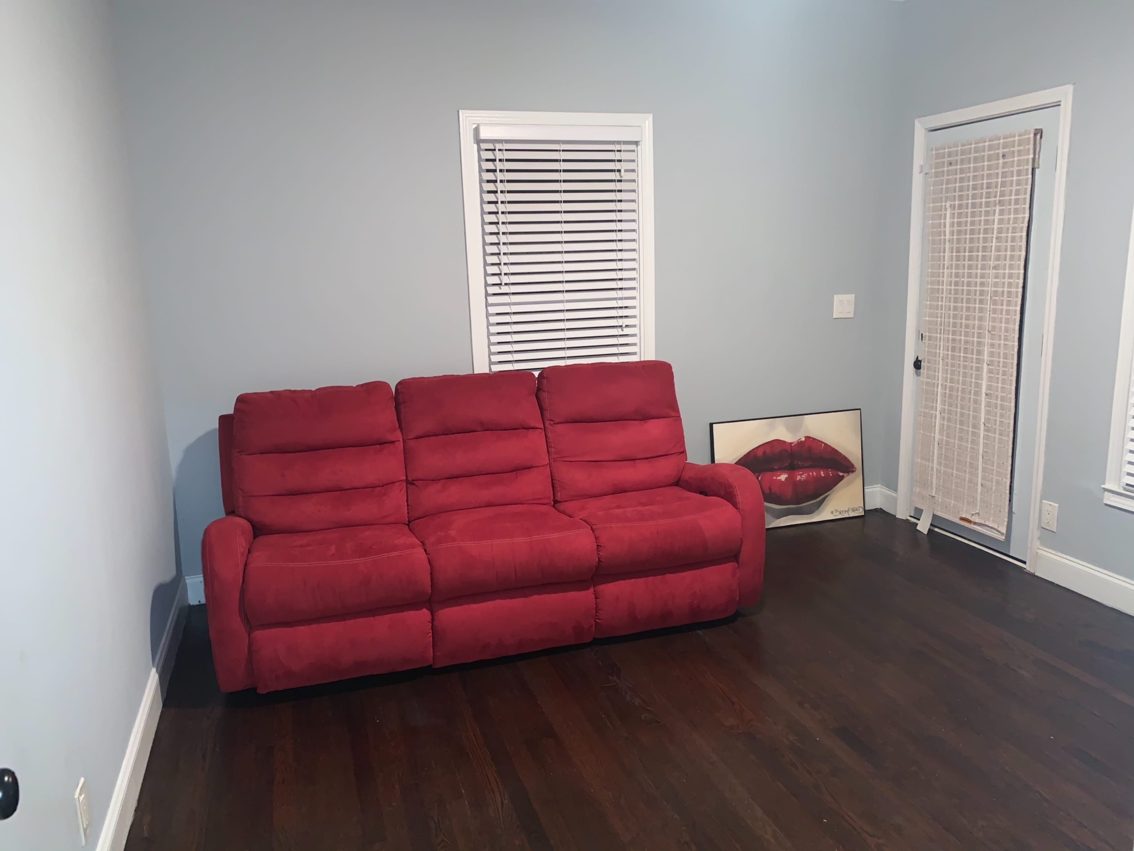 A red couch is positioned against a blue wall in a sparsely furnished room with a door and a framed artwork on the floor.