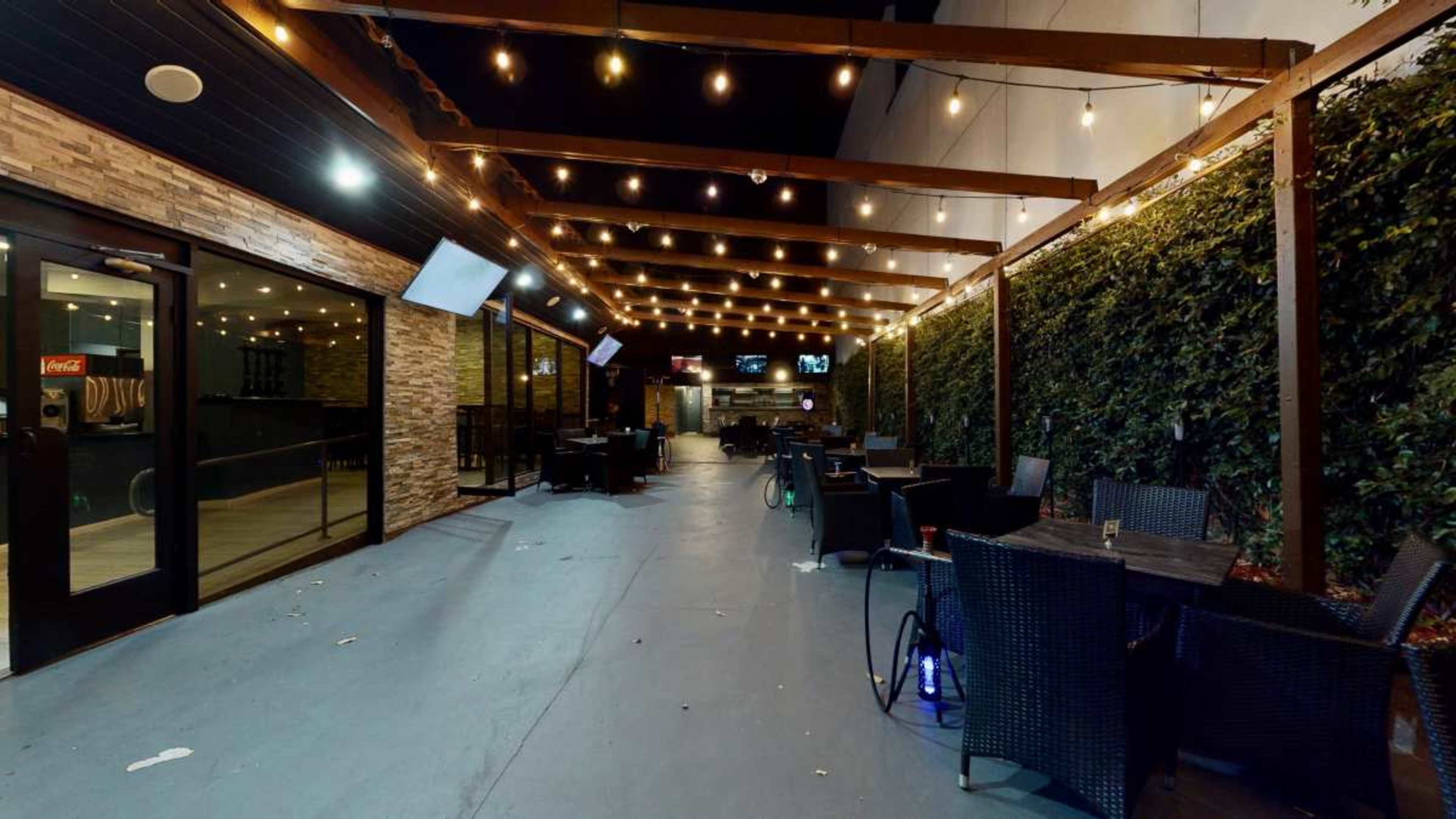 The image shows an outdoor dining area with string lights overhead and several tables and chairs arranged along a pathway.