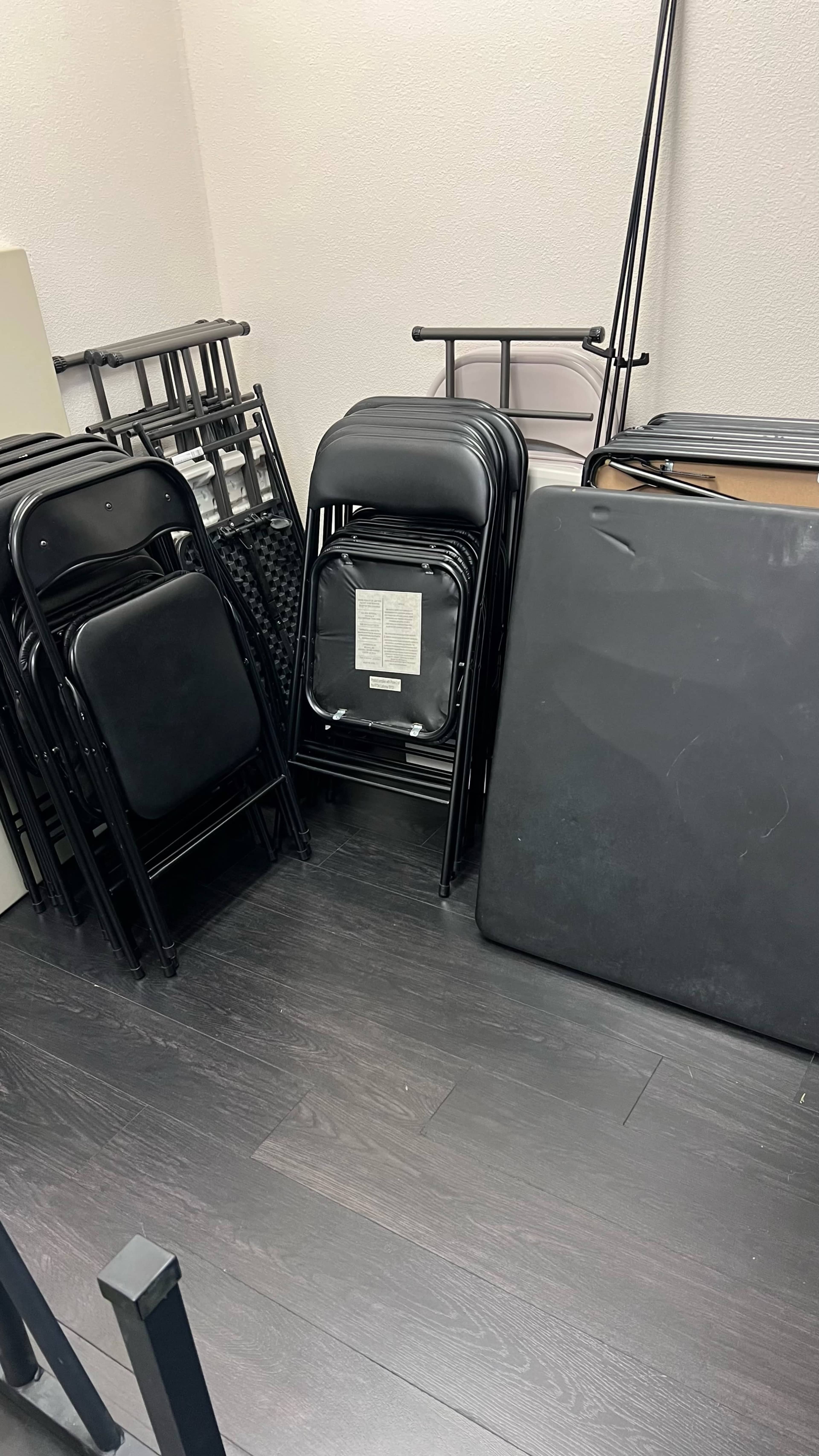 A stack of black folding chairs is positioned next to a flat black table against a wall in a small room.