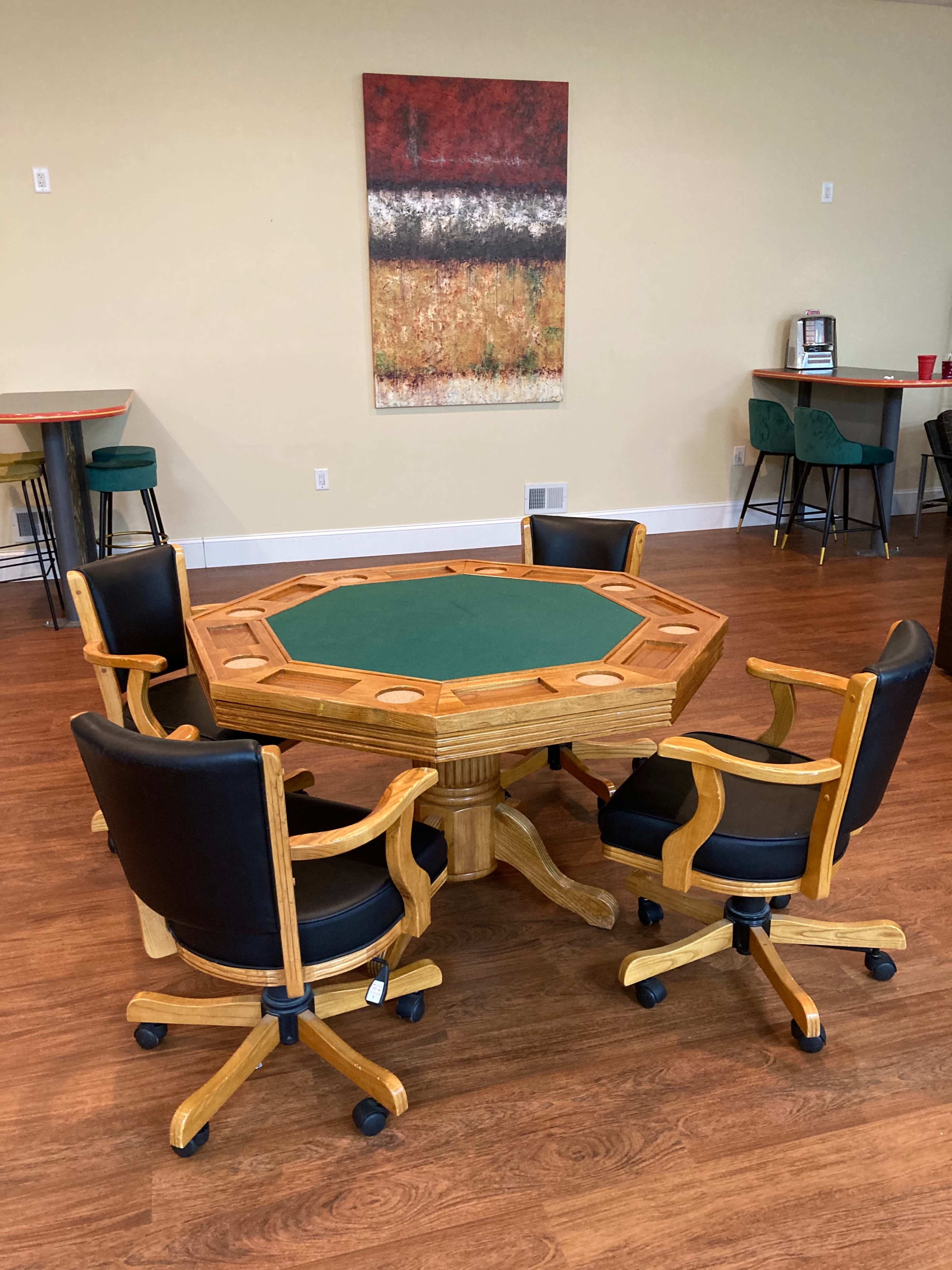 A wooden octagonal poker table with a green felt surface is surrounded by four black swivel chairs in a room with a simple wall decor.