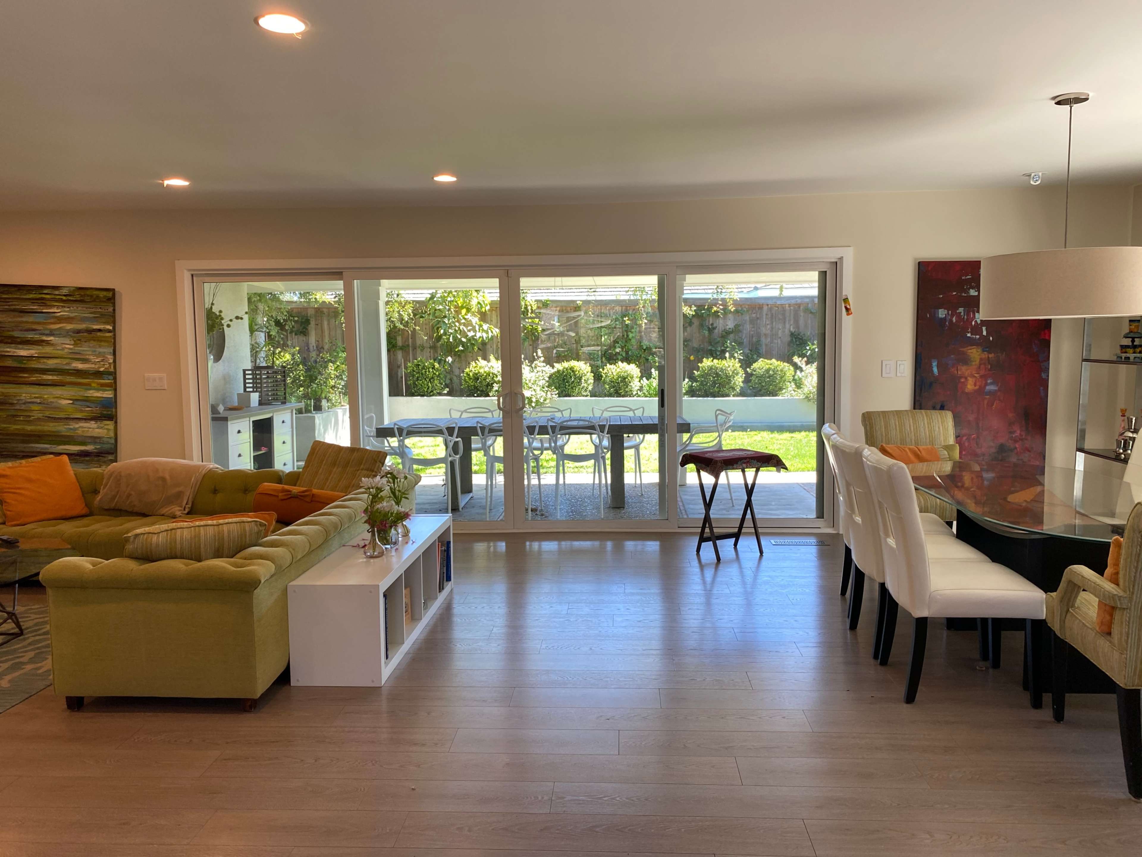 A bright living area features a green sectional sofa, a dining table with white chairs, and large windows overlooking a garden.