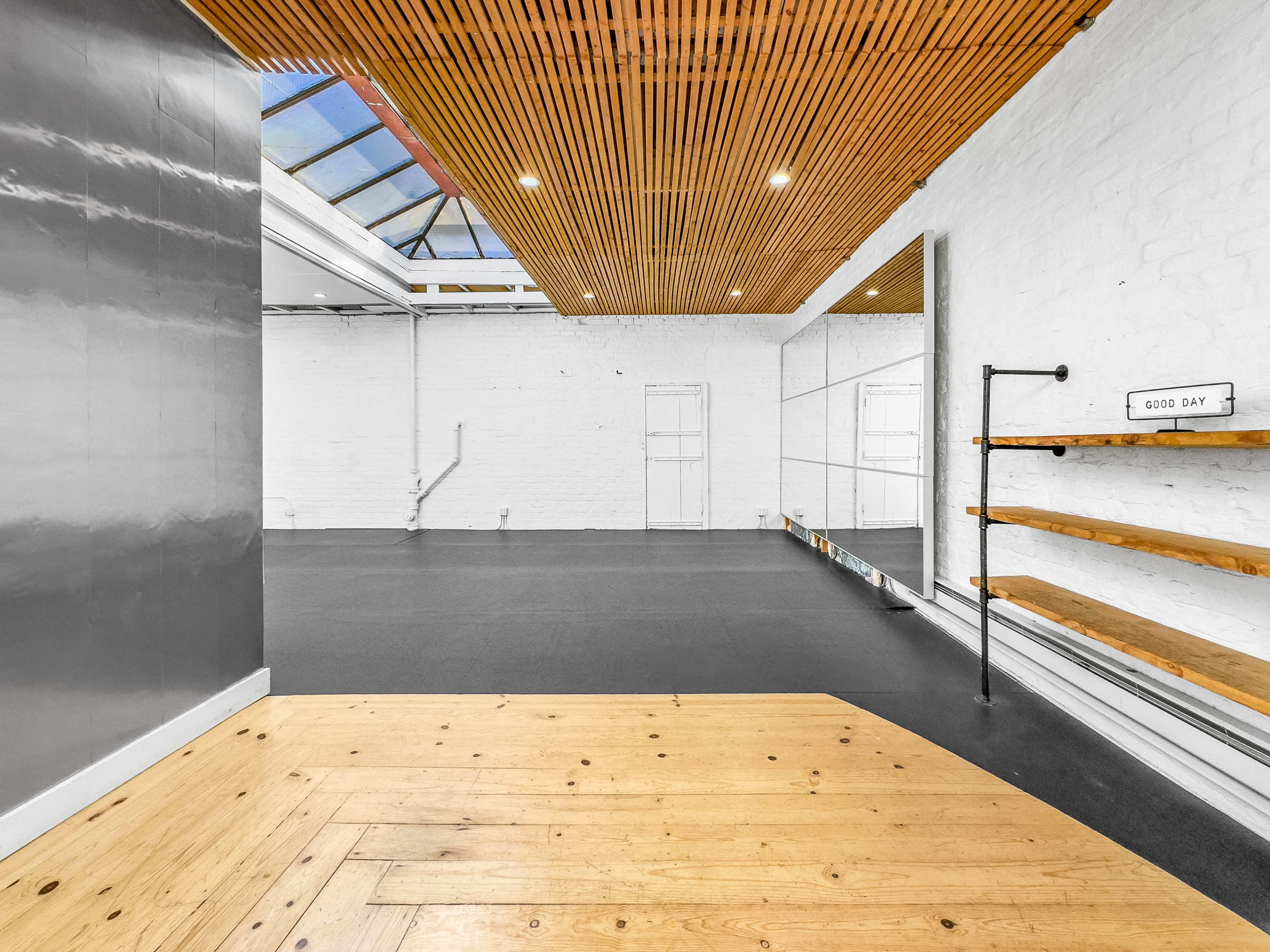 A spacious studio area with wooden flooring, a mirrored wall, and wooden shelving against a white brick backdrop.