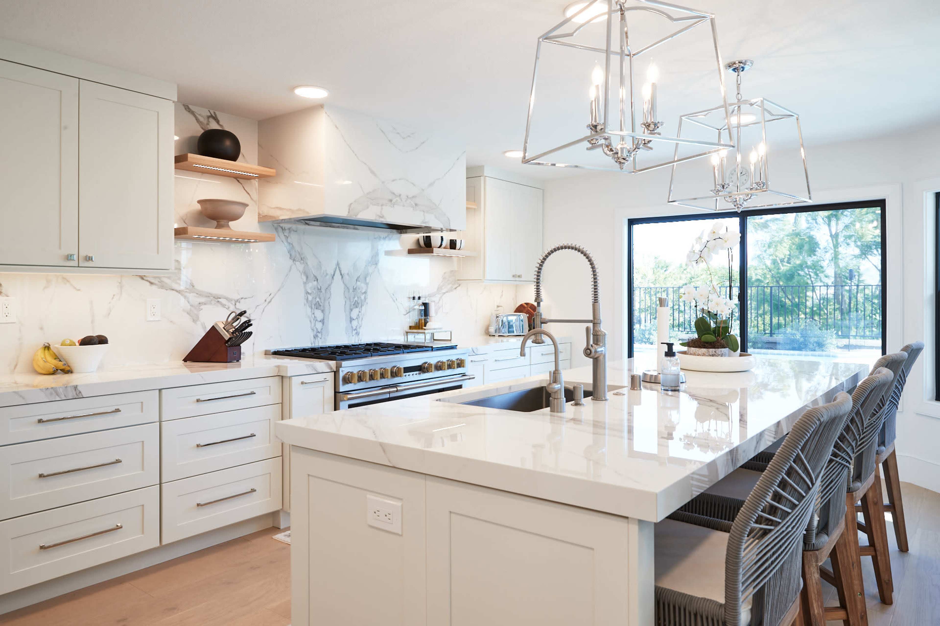 A modern kitchen featuring white cabinetry, a large marble island with bar stools, and a sliding glass door leading to an outdoor area.