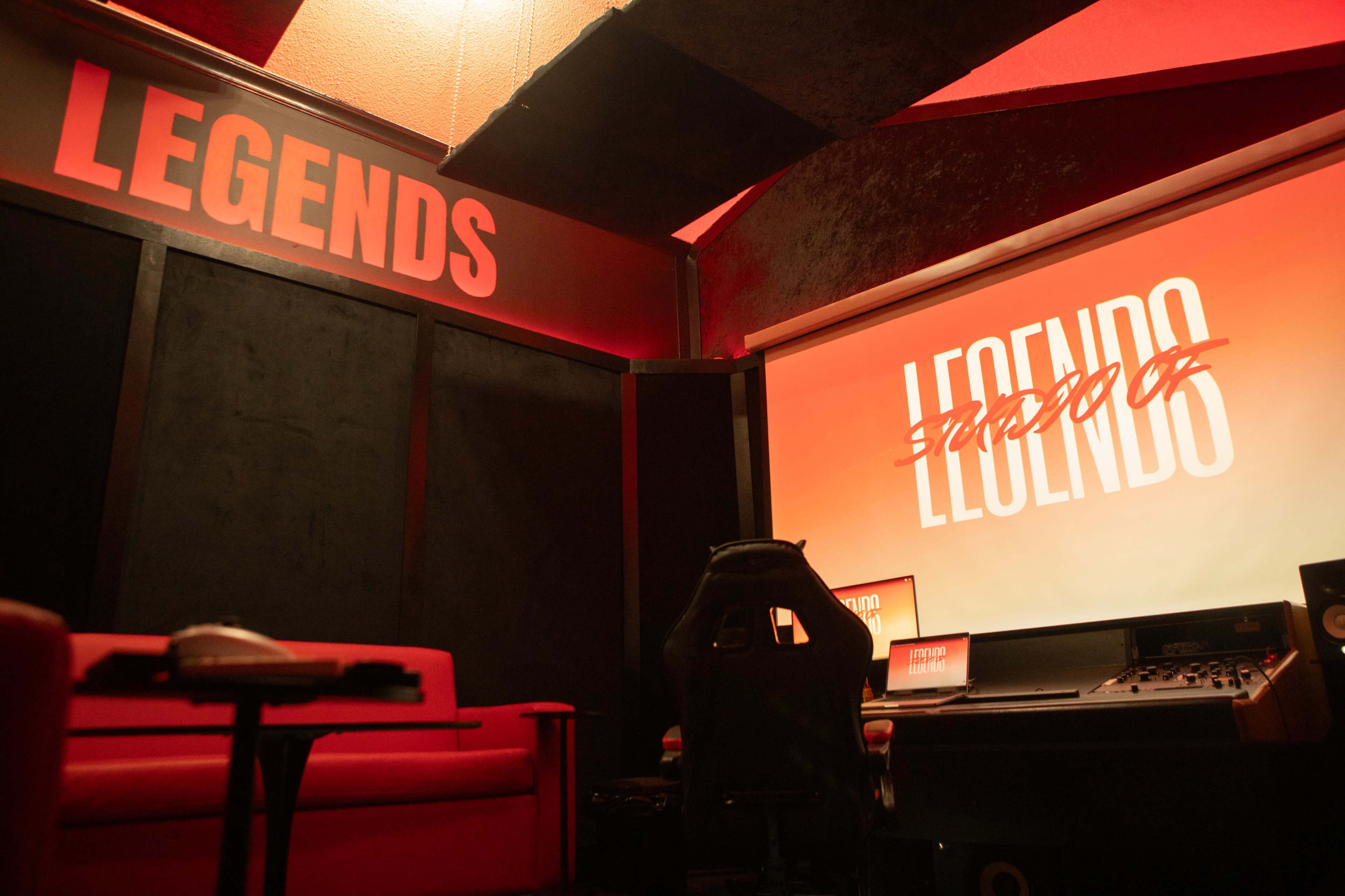 The image shows a dimly lit room designed for creative work, featuring a large screen displaying the word "LEGENDS" and a red couch in front of a sound mixing console.