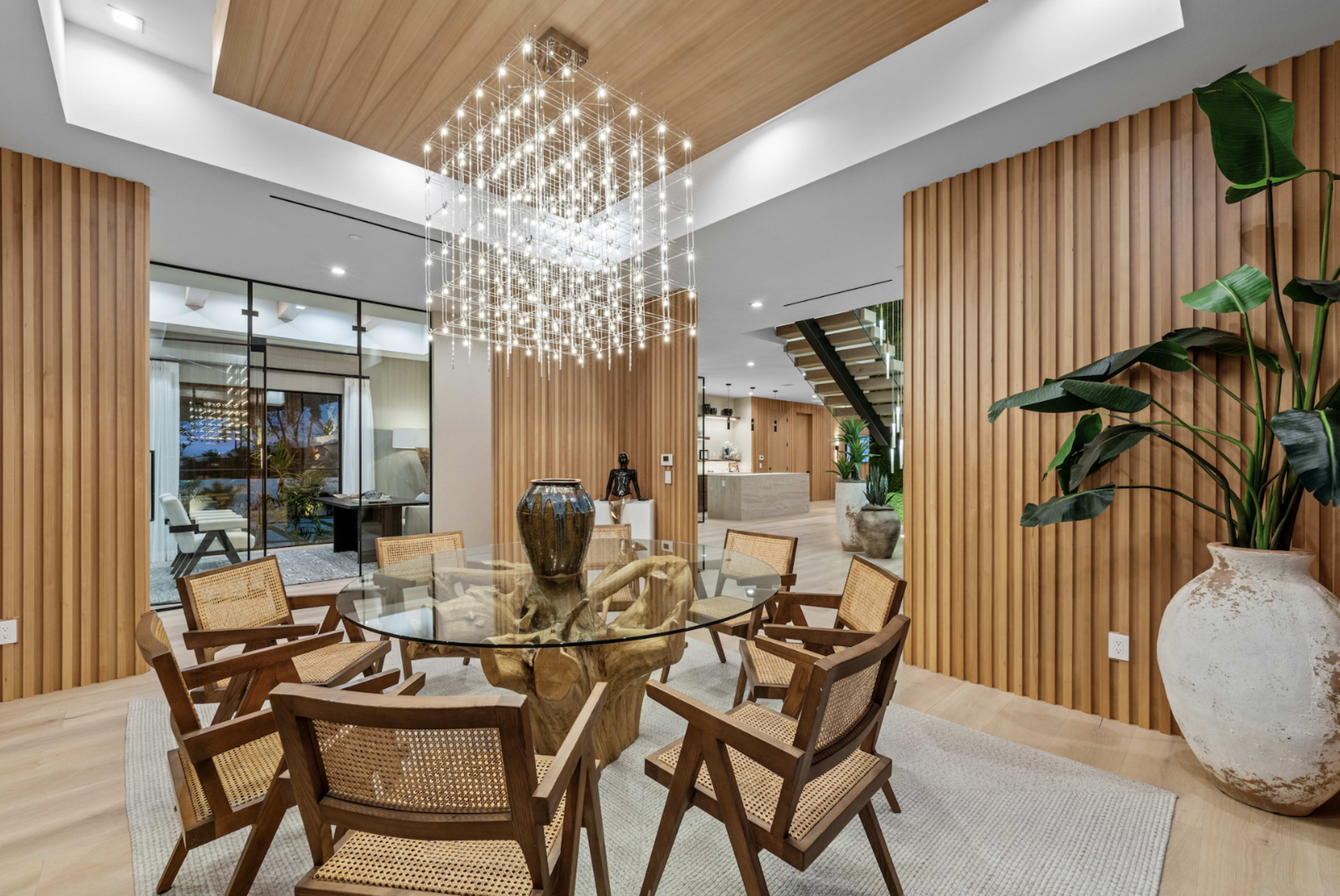 The image shows a contemporary dining area featuring a round glass table surrounded by wooden chairs, with a chandelier above and wooden paneling on the walls.