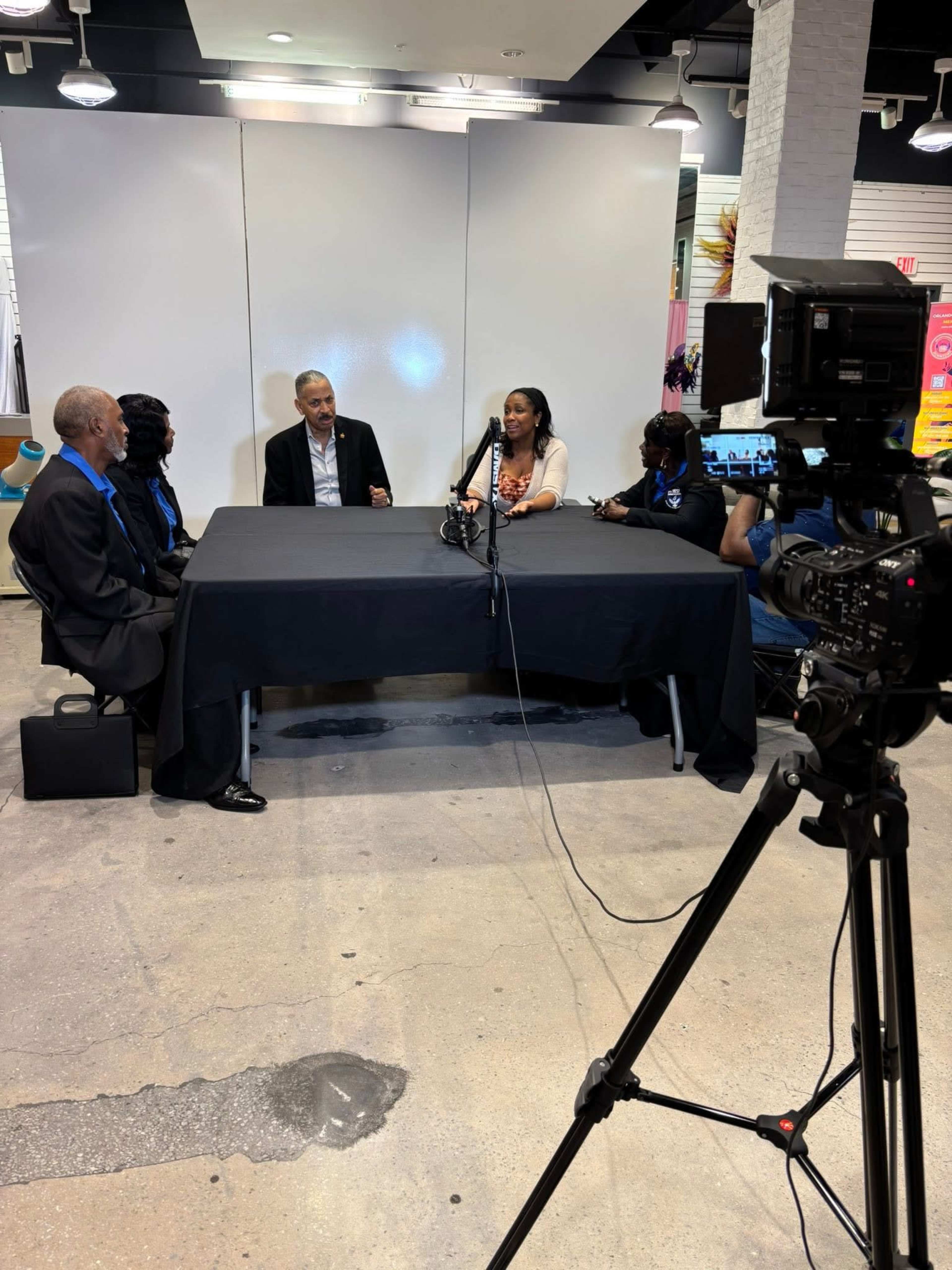 A group of people is seated at a table, engaged in discussion, while a camera is positioned nearby capturing the scene.