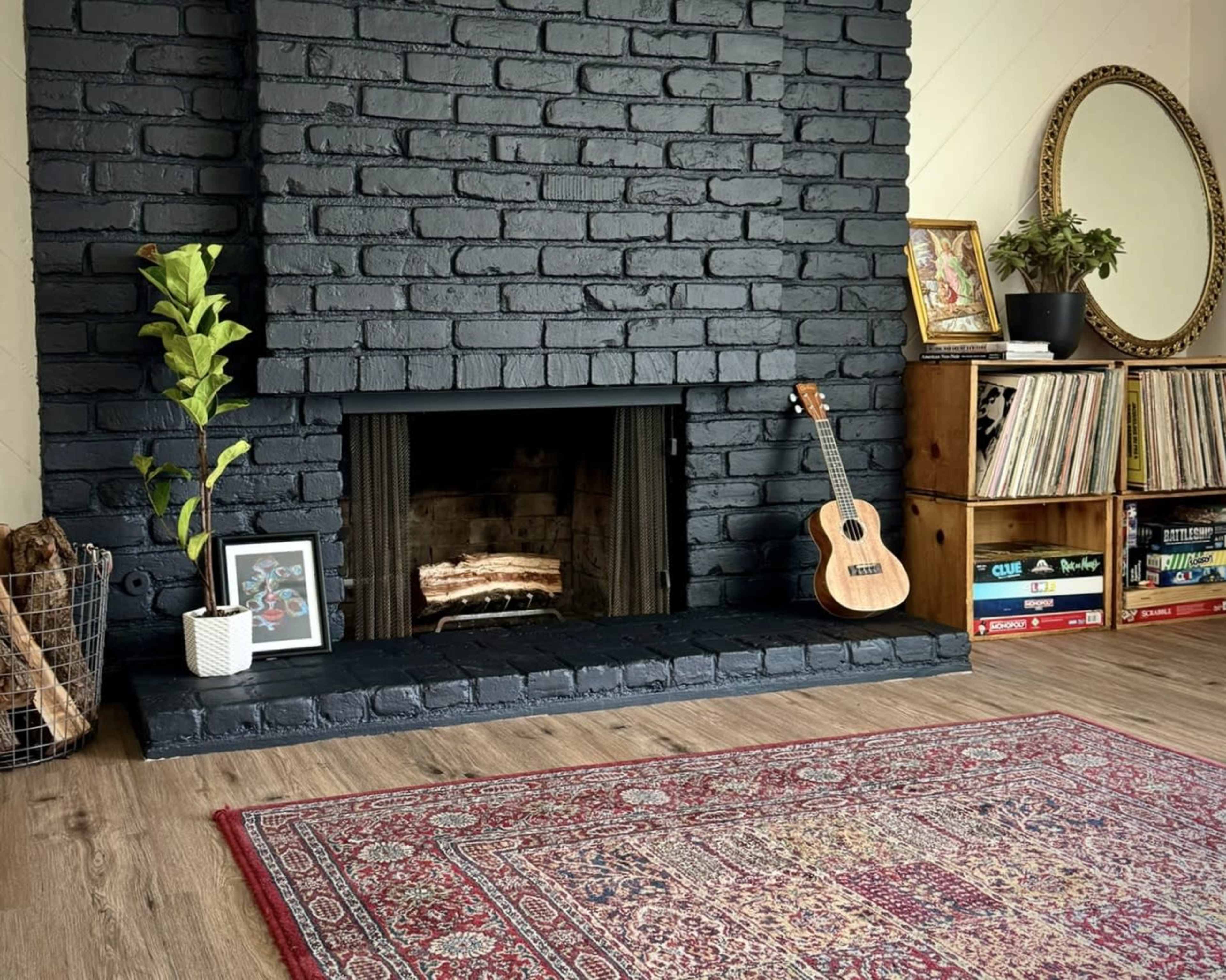 A black brick fireplace is the focal point of a living room, flanked by a guitar, a decorative mirror, and a wooden bookshelf filled with books.
