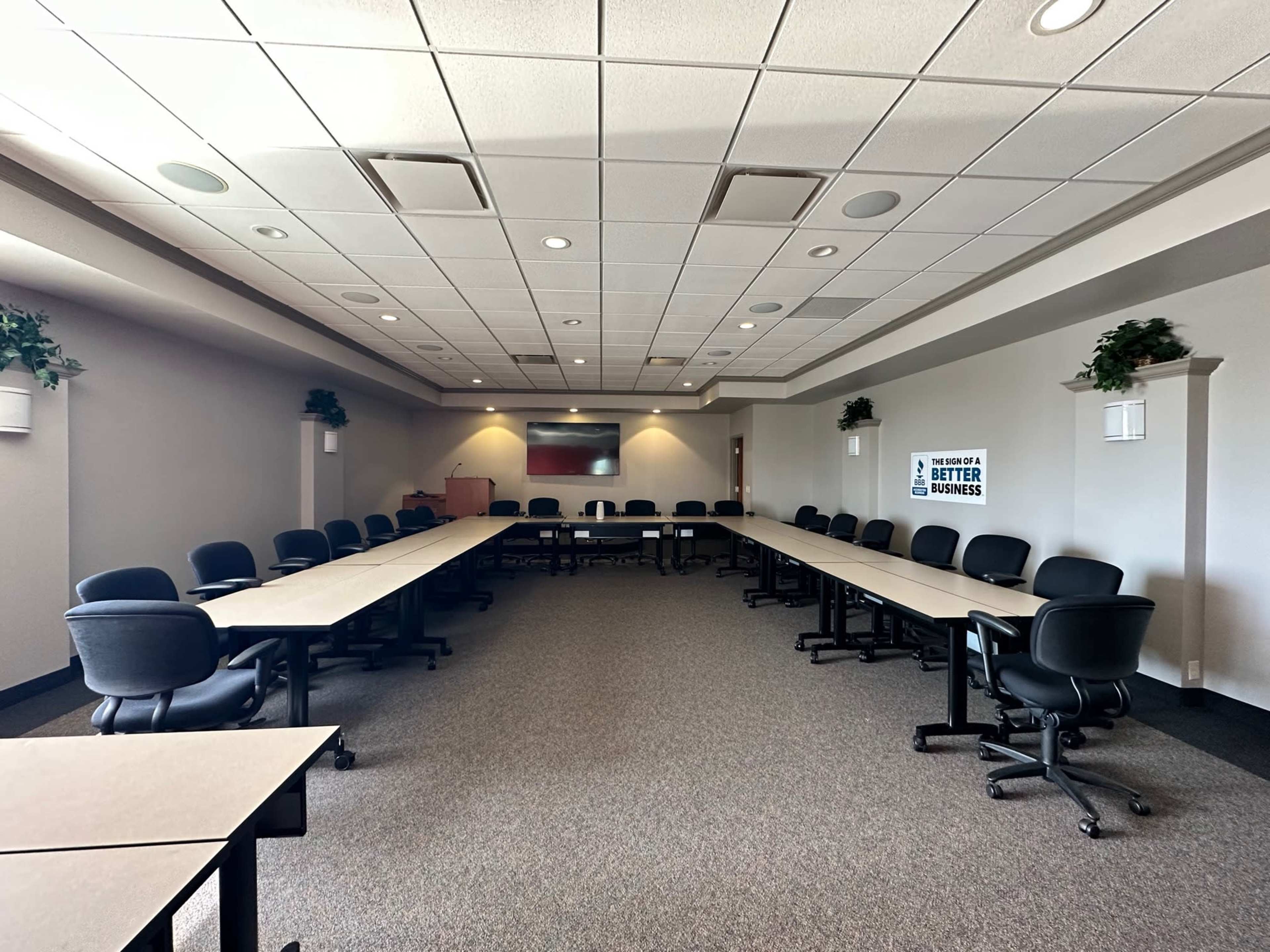 A spacious conference room features a U-shaped arrangement of tables with black chairs and a presentation area at the front.