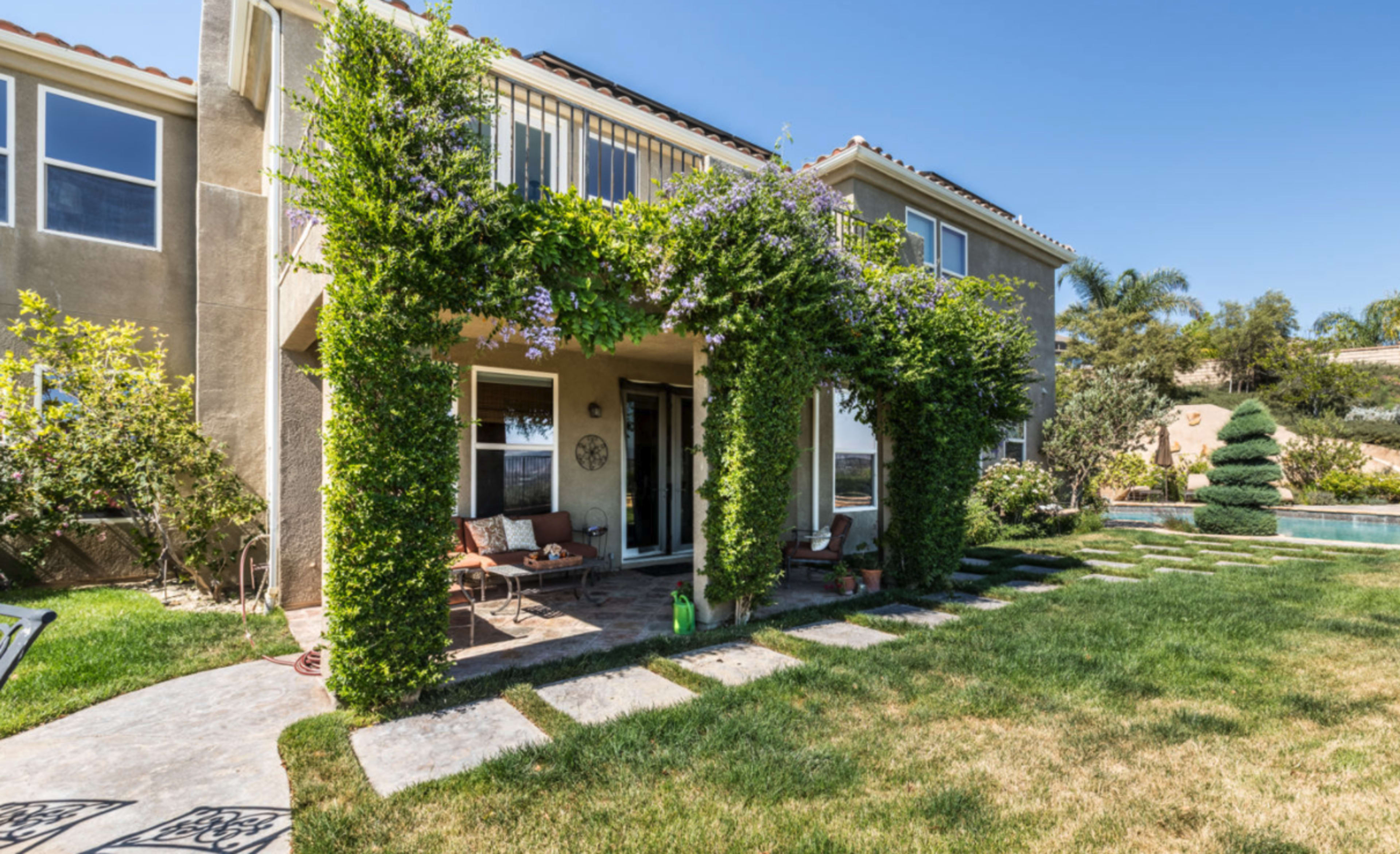 The image shows a two-story house with a patio covered in climbing vines and a lawn leading to a swimming pool in the background.