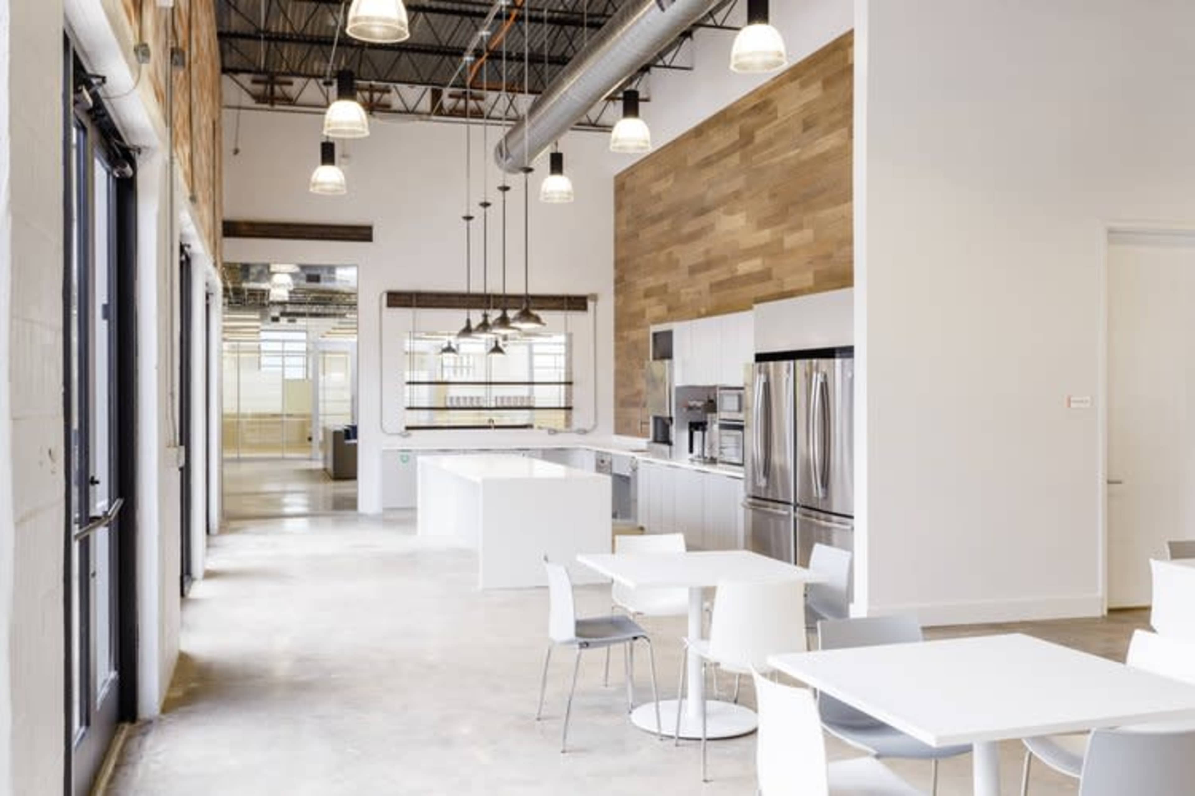 A modern kitchen and dining area with white tables and chairs, a large island, and stainless steel appliances against a backdrop of wood paneling.