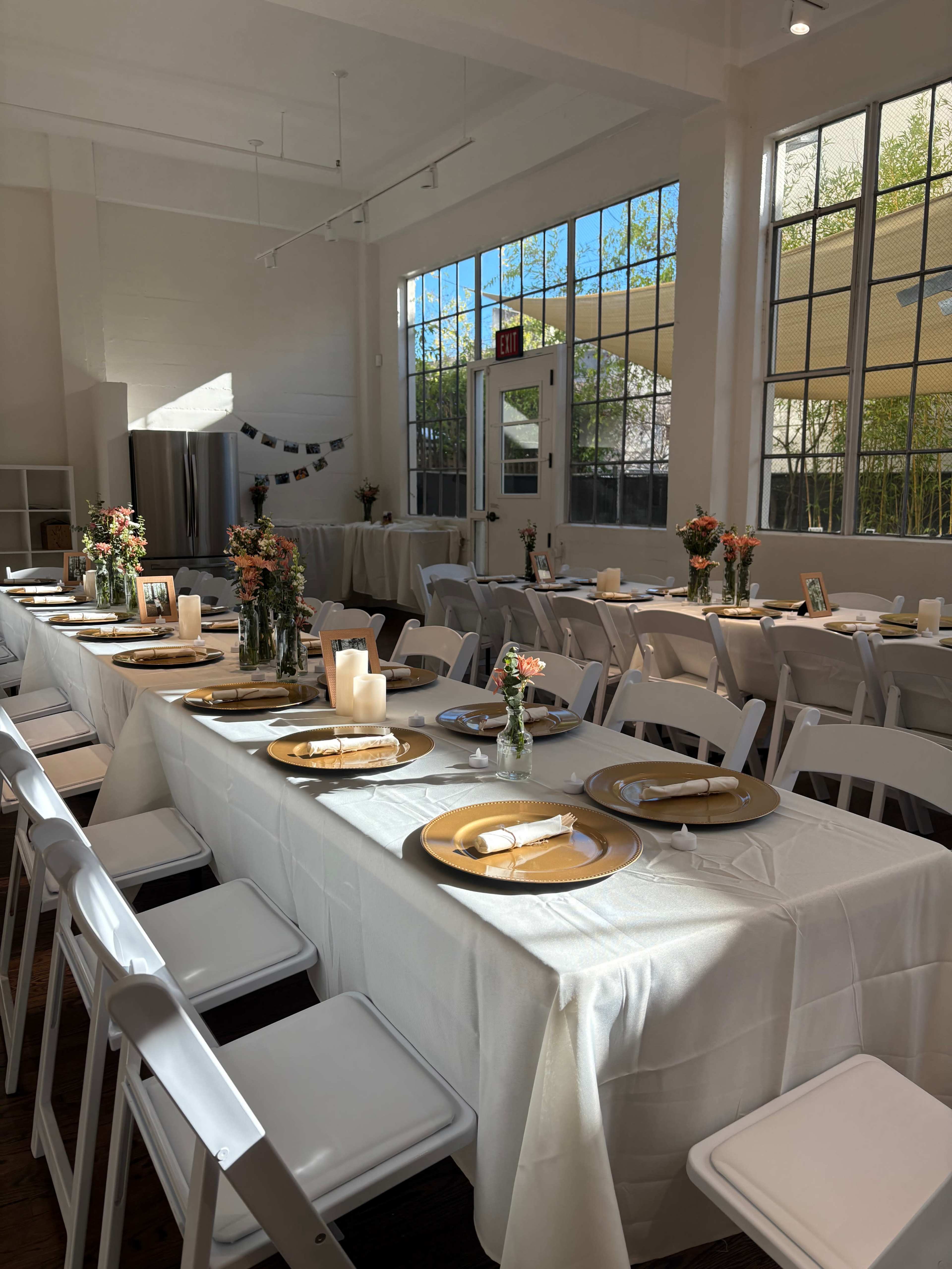 A spacious indoor event space features neatly arranged tables with white tablecloths, gold plates, and floral centerpieces, illuminated by sunlight streaming through large windows.