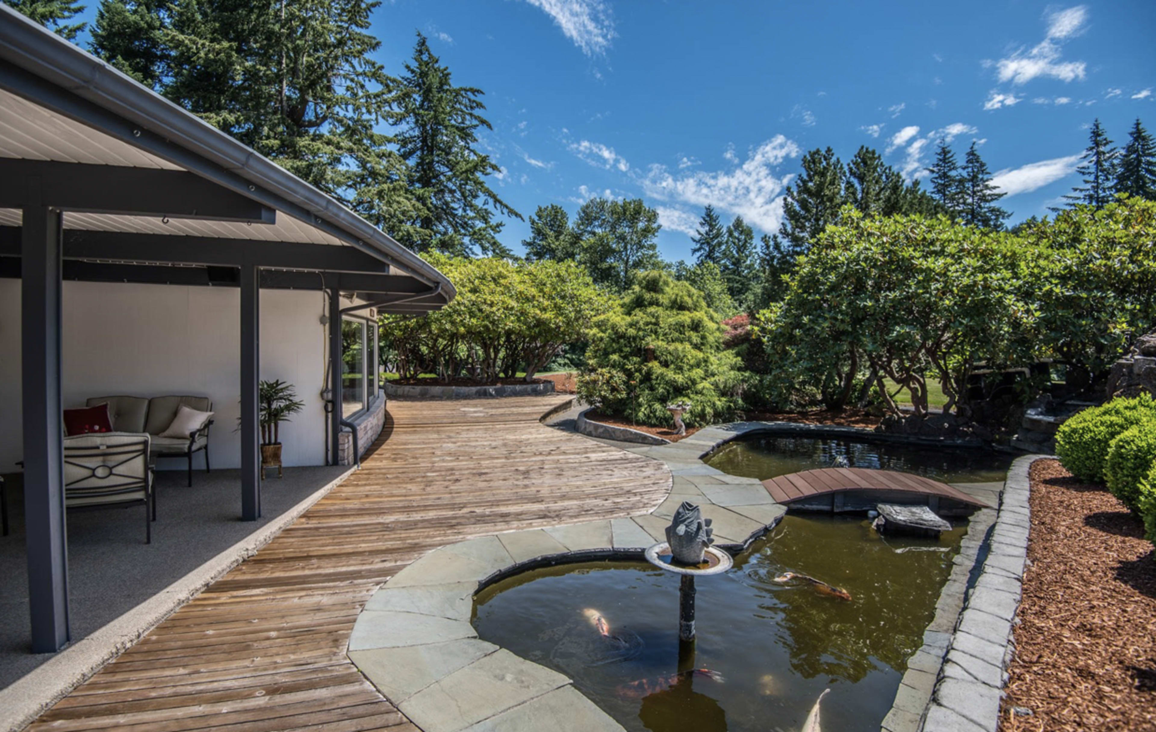 A landscaped backyard with a wooden deck, a koi pond, and a small arched bridge surrounded by greenery.