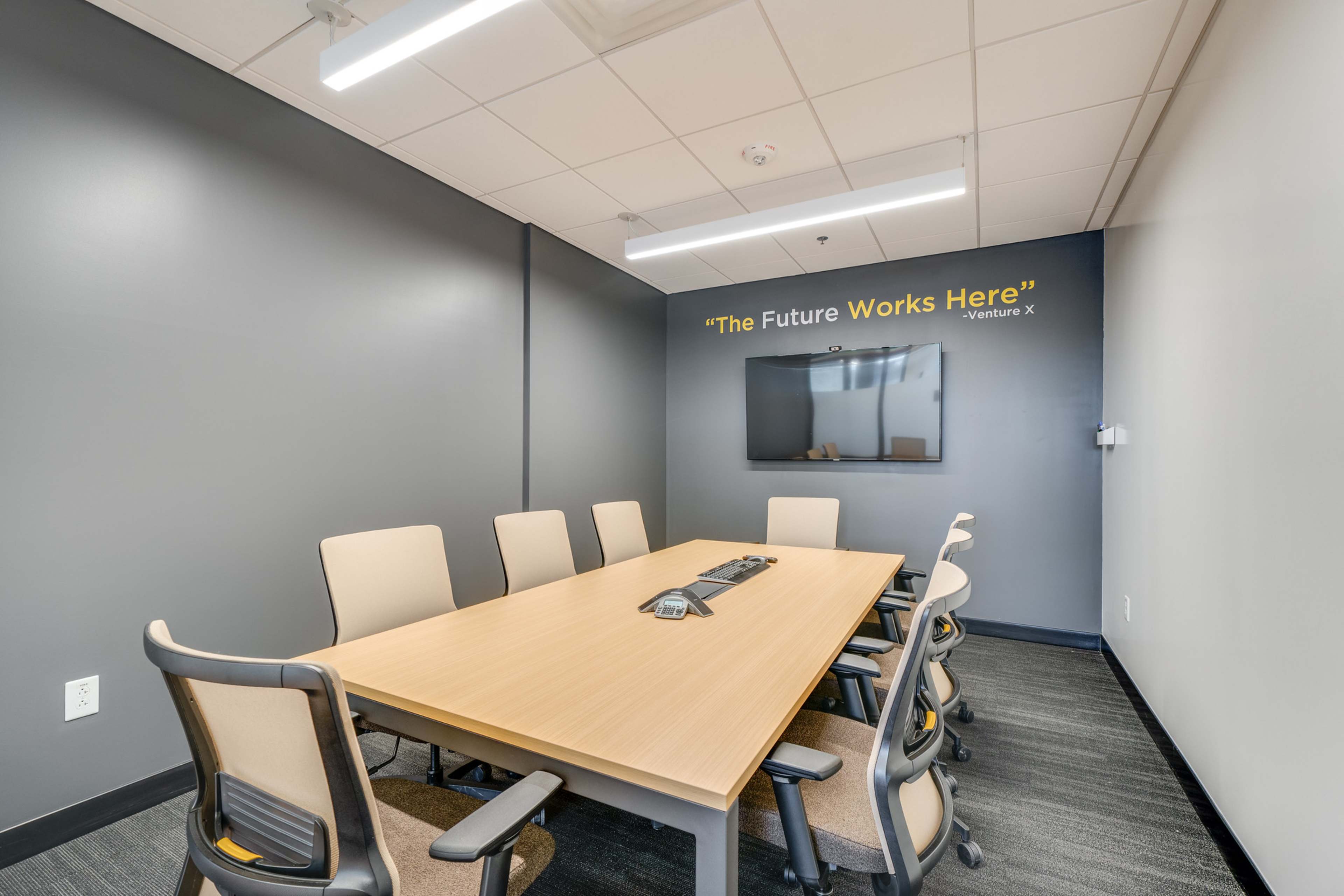 A conference room features a rectangular table surrounded by eight chairs, with a wall-mounted screen and a motivational quote above it.