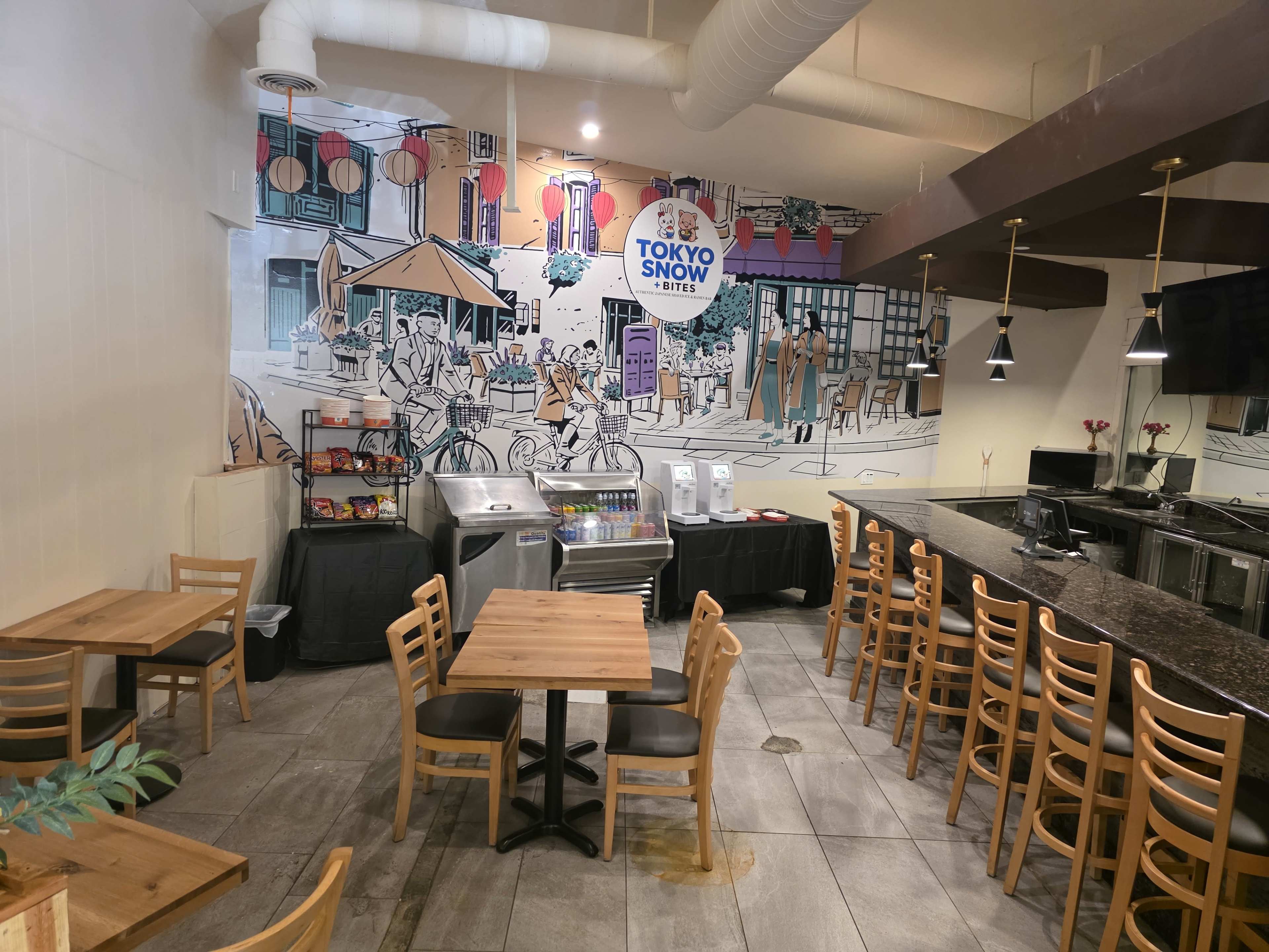 The image shows the interior of a café featuring wooden tables, chairs, and a mural of a city scene on the wall, along with a service counter and drink dispensers.
