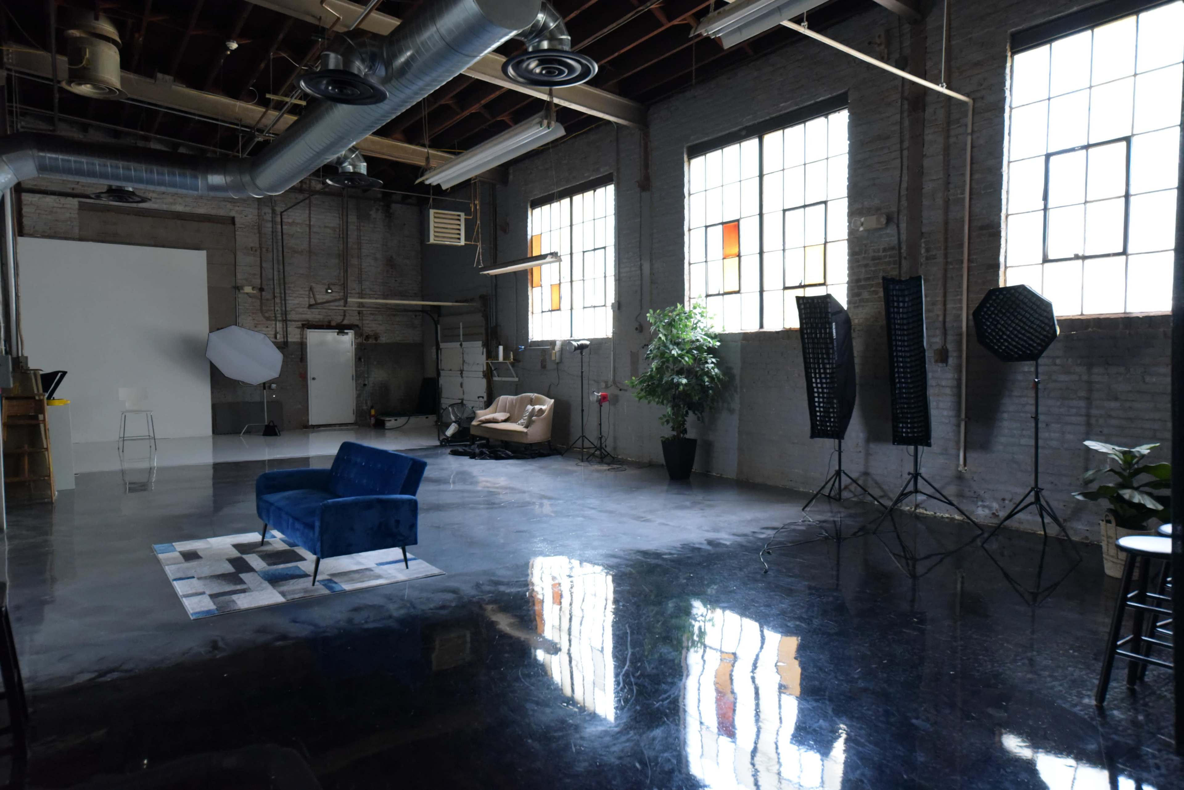 A spacious studio with polished concrete floors, large windows, various lighting equipment, and two sofas placed against one wall.
