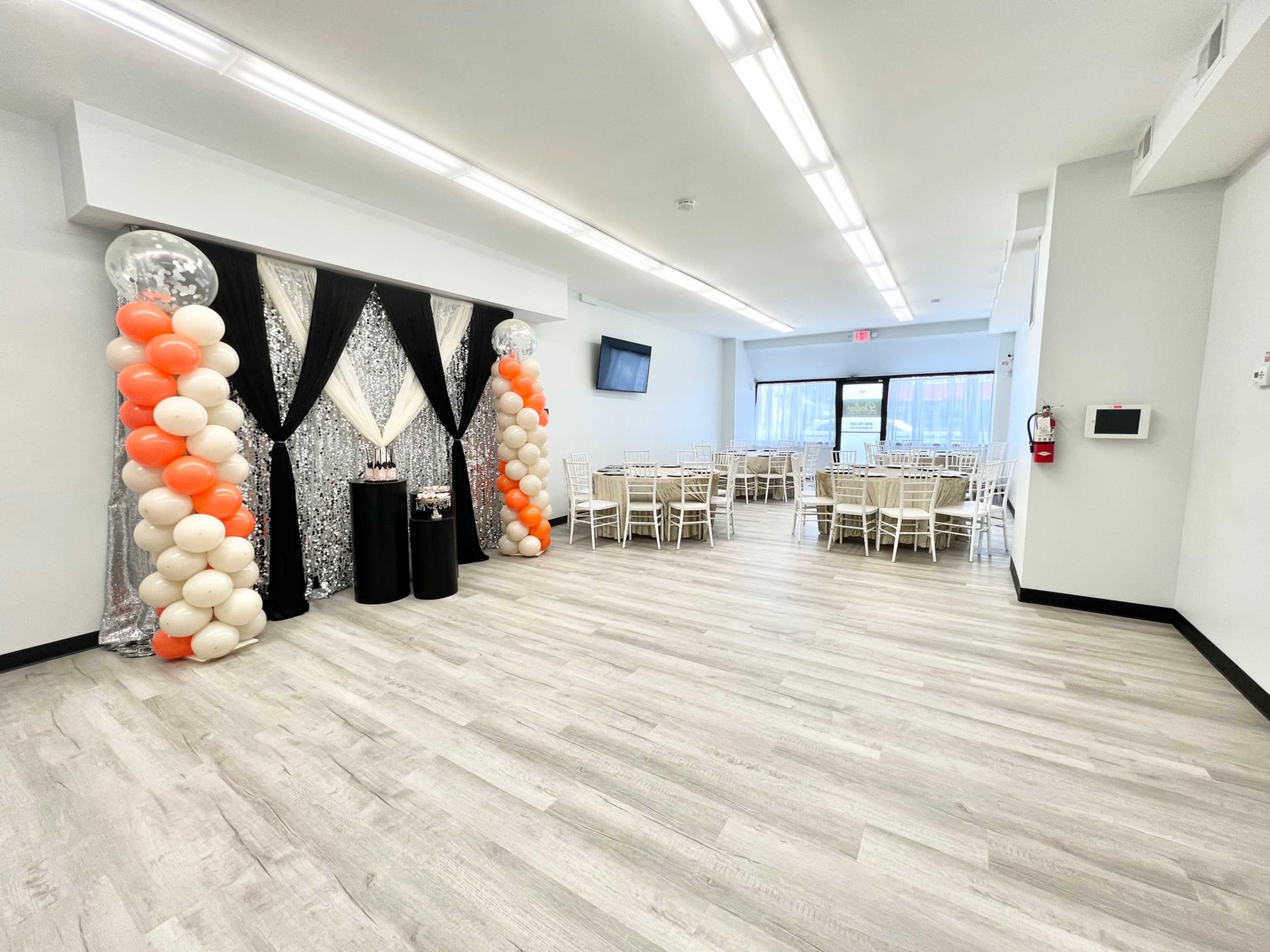 The image shows a bright, modern event space featuring a decorated entrance with balloons and two tables set up for a gathering, while several round tables with chairs are arranged in the background.