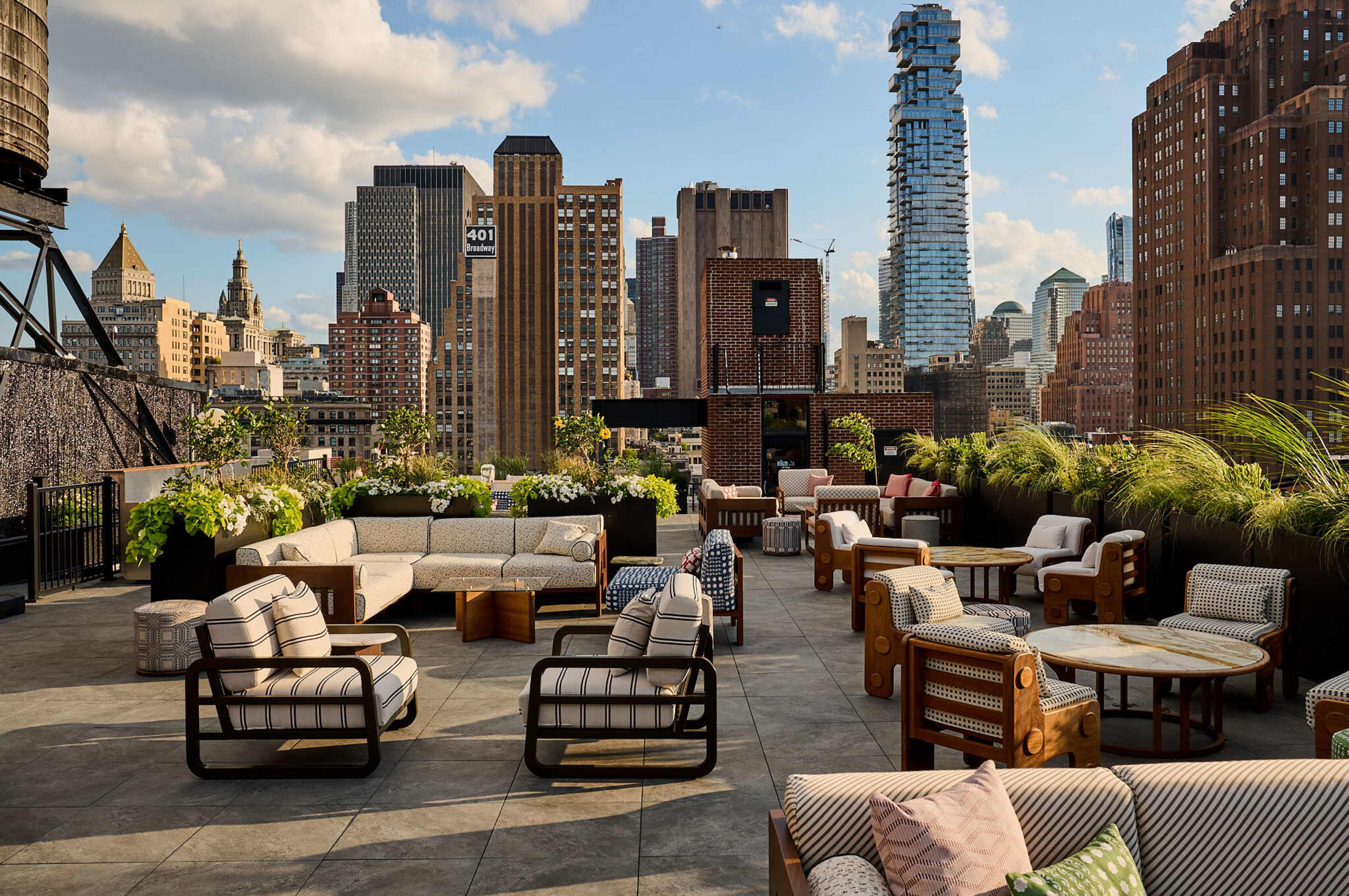 The image shows a rooftop patio with modern furniture, lush greenery, and a skyline view of tall buildings under a partly cloudy sky.