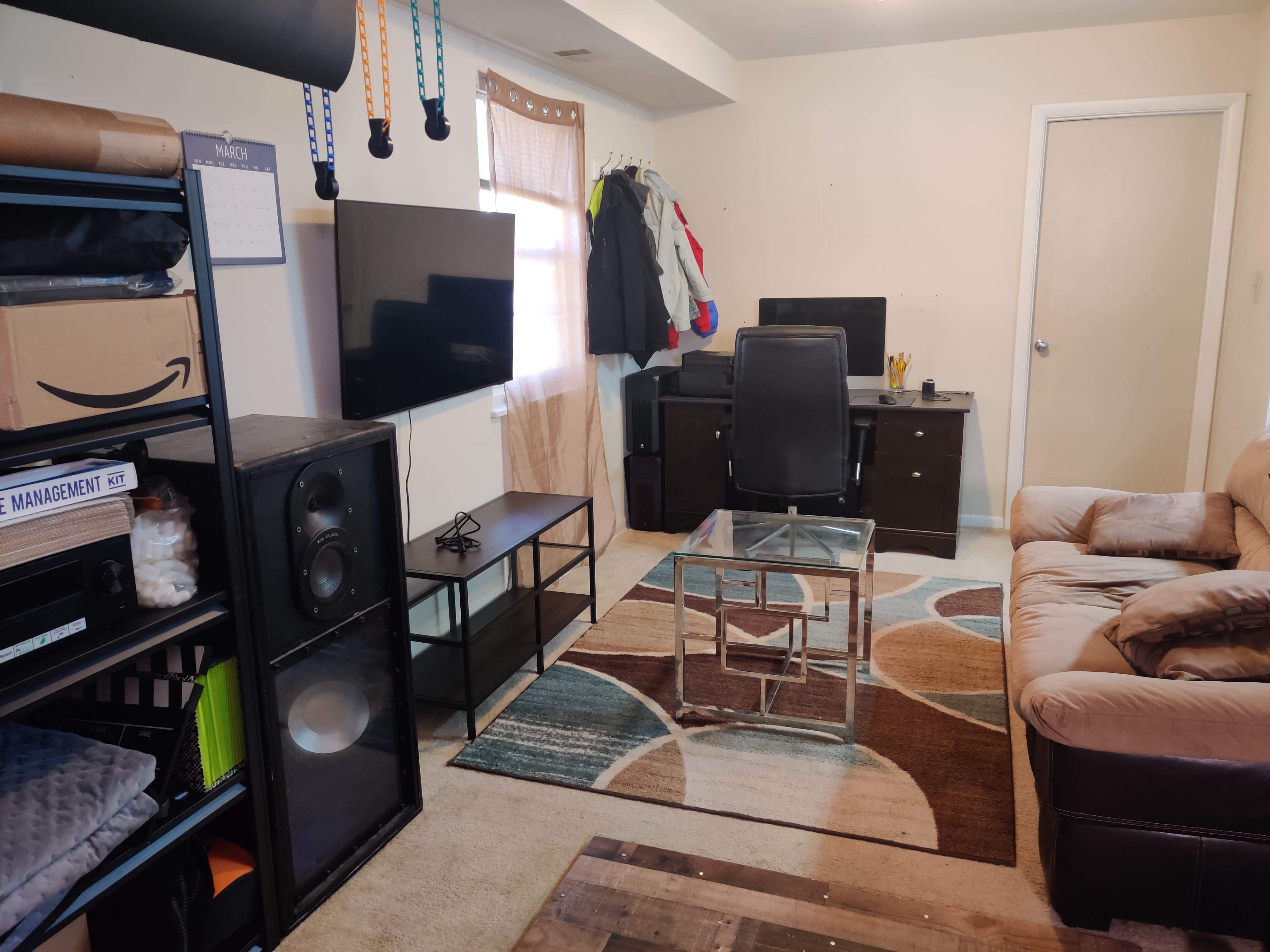 The image shows a small room featuring a couch, a glass coffee table, a desk with a computer setup, and a TV, along with storage shelves and a rug on the floor.