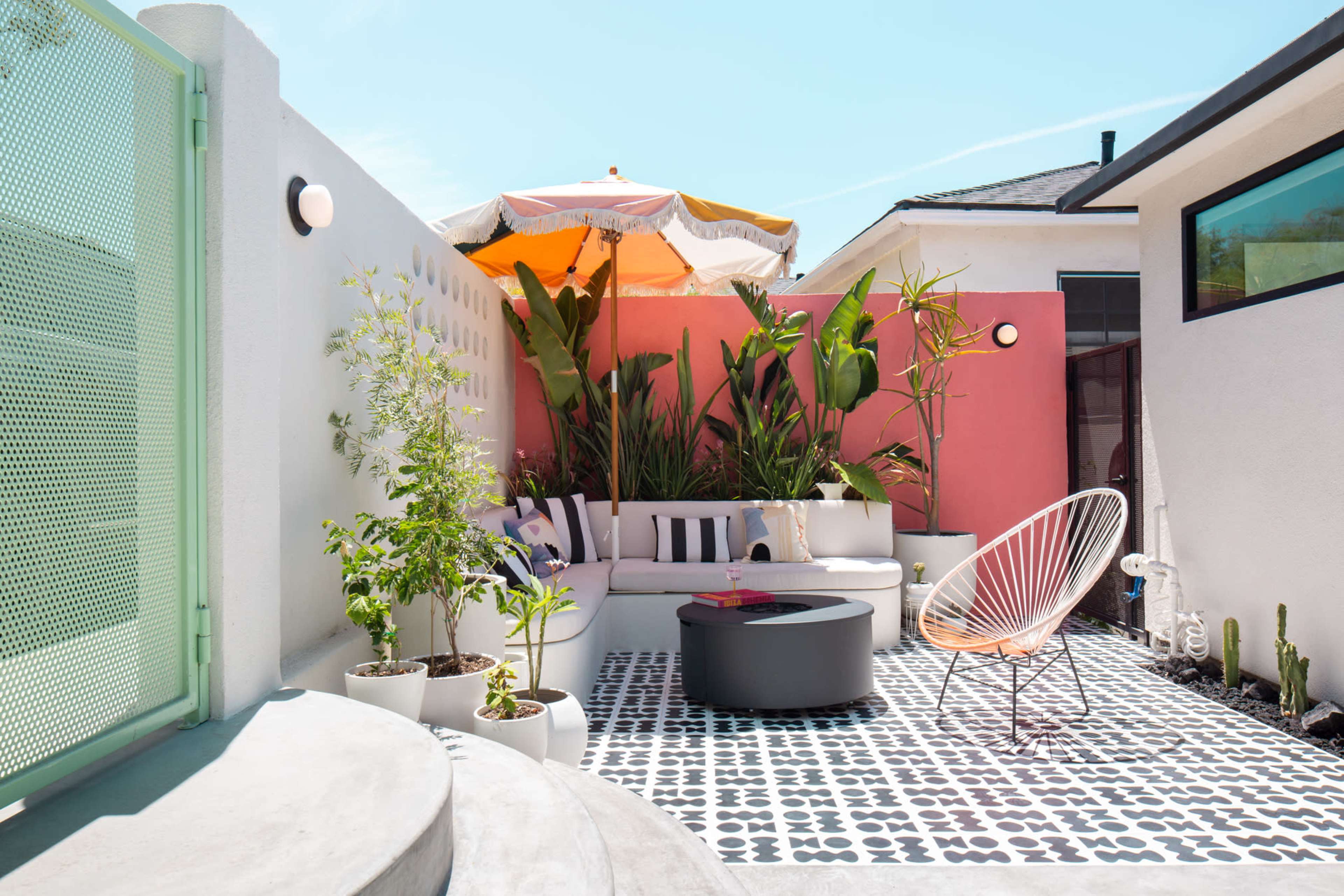 The image shows a bright outdoor patio area with a modern seating arrangement, potted plants, and a colorful umbrella providing shade.