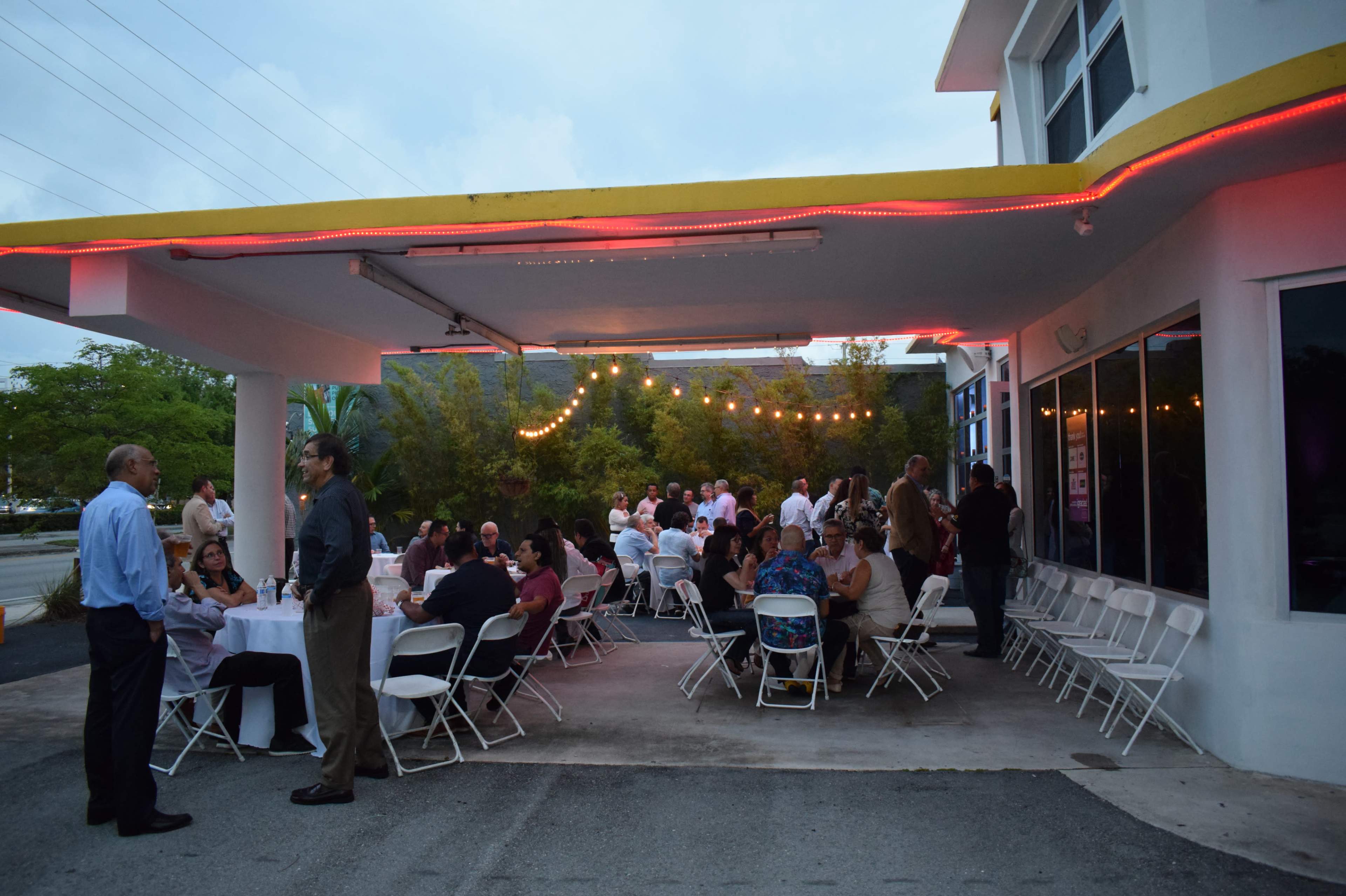 A group of people is gathering under a covered outdoor area, with tables set up for dining and string lights illuminating the scene.
