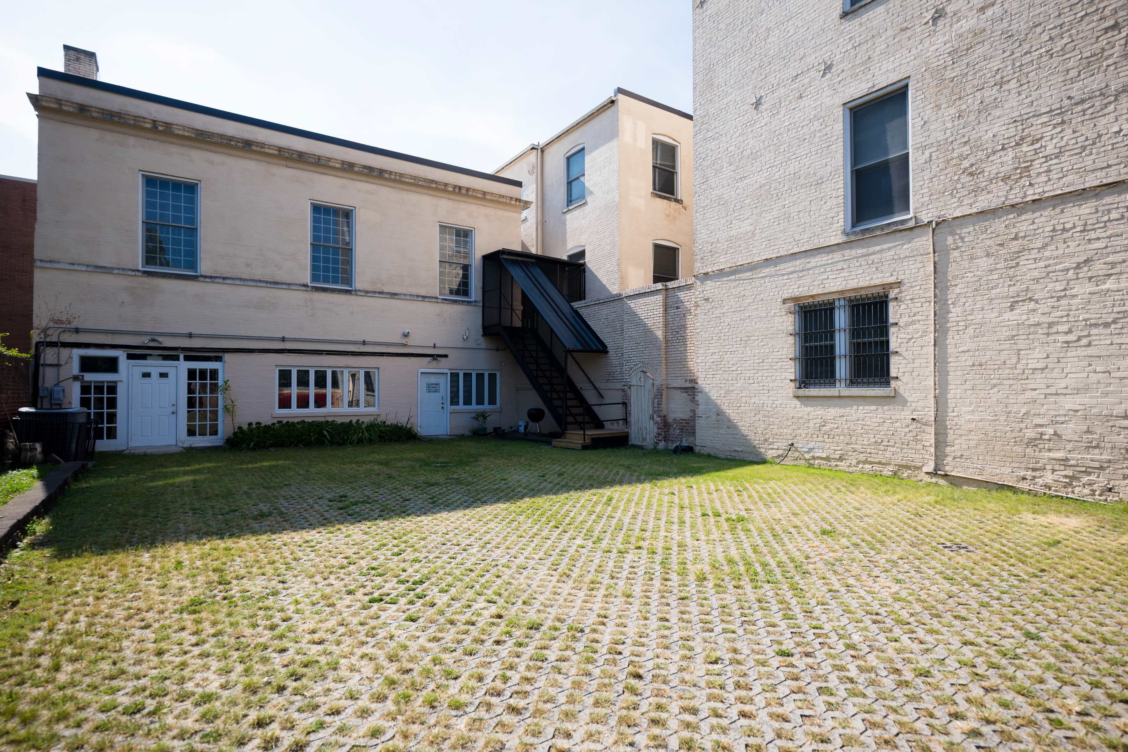 The image shows a vacant courtyard surrounded by two buildings, with stairs leading to an upper level and a pattern of grass and paving stones on the ground.