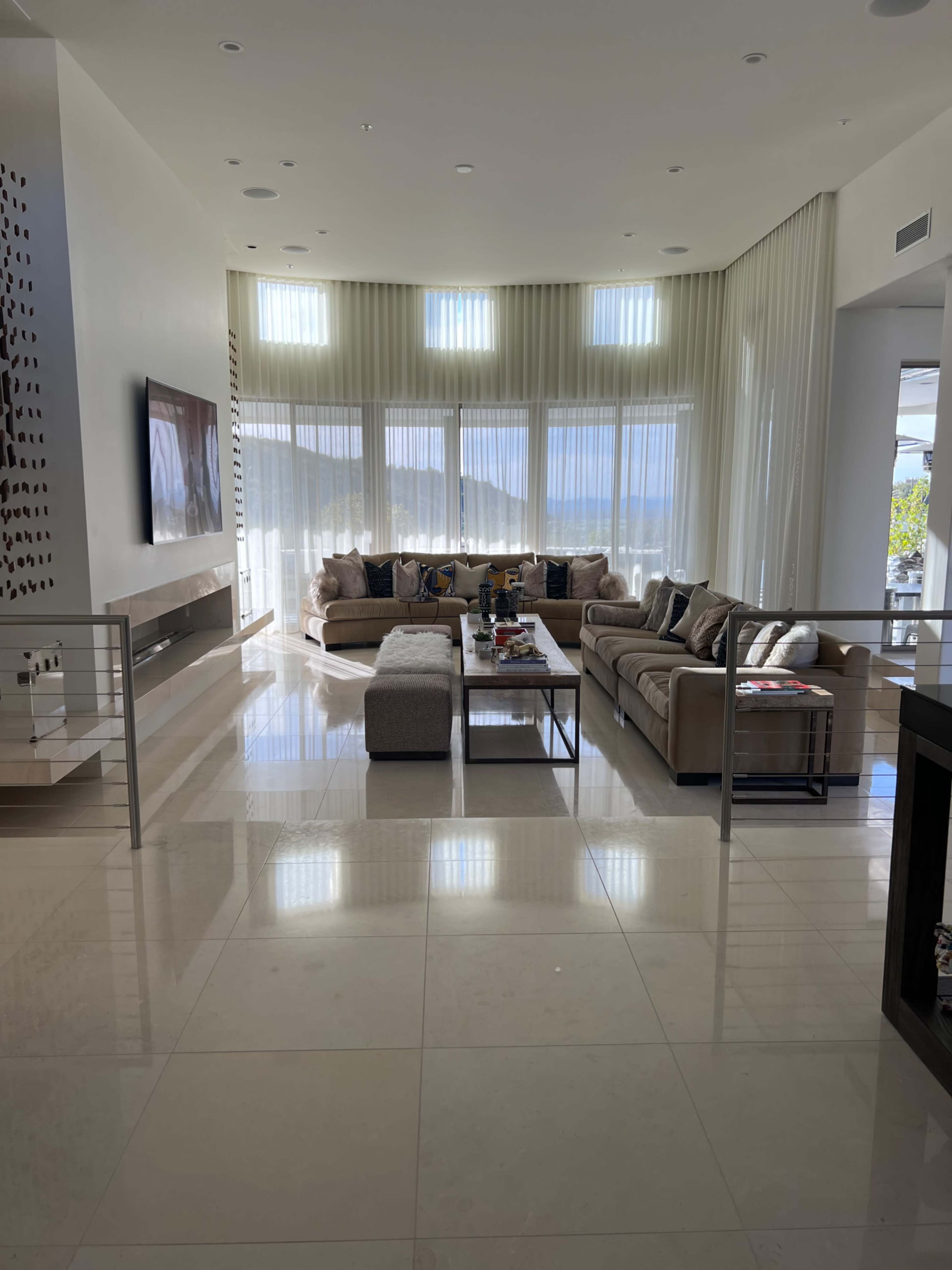 The image shows a modern living room with large windows, a beige sectional sofa, and a coffee table situated on a polished tile floor.