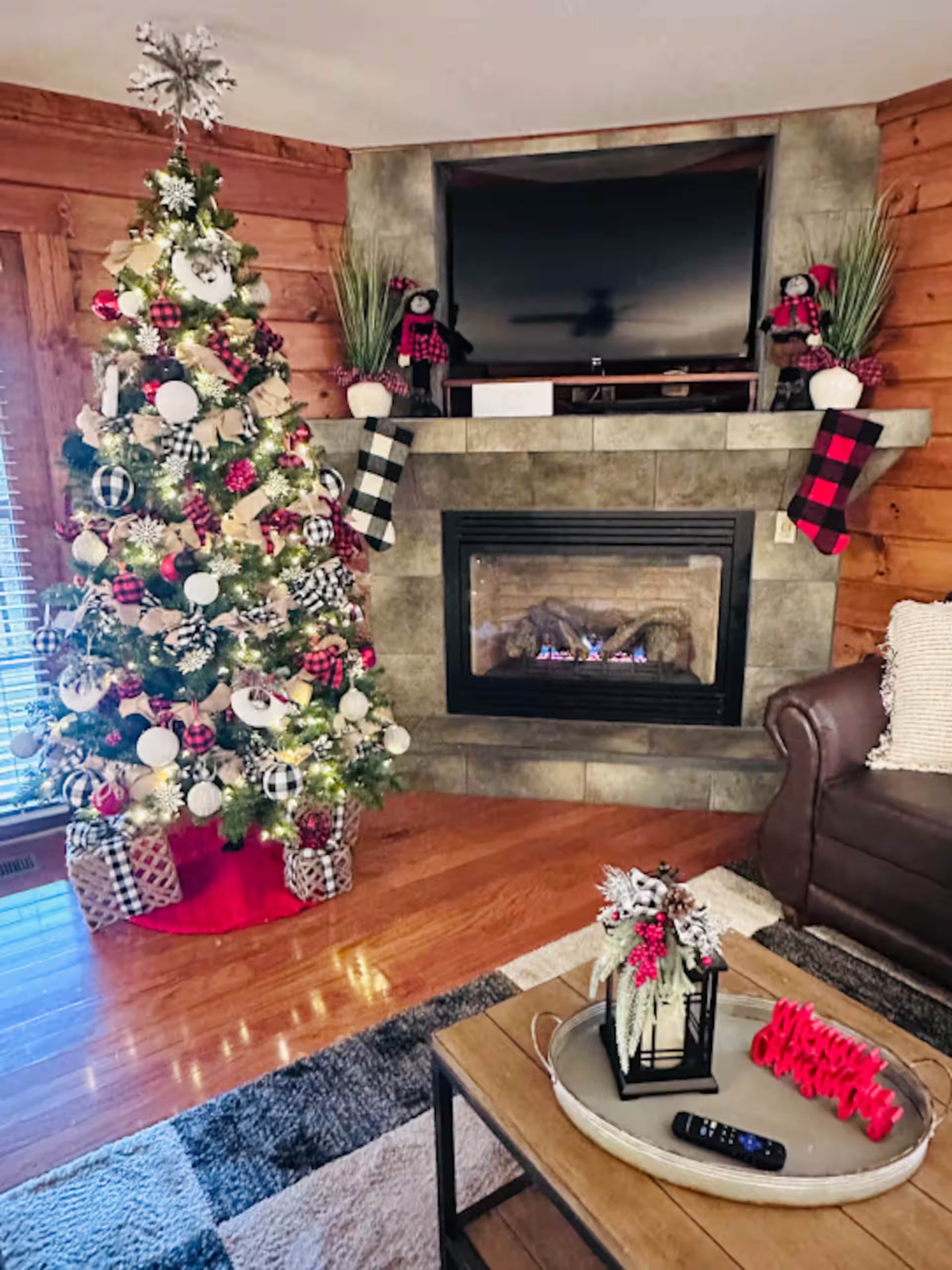 The image shows a cozy living room decorated for the holidays, featuring a Christmas tree with ornaments, a stone fireplace with stockings, and a wooden coffee table with decorative items.