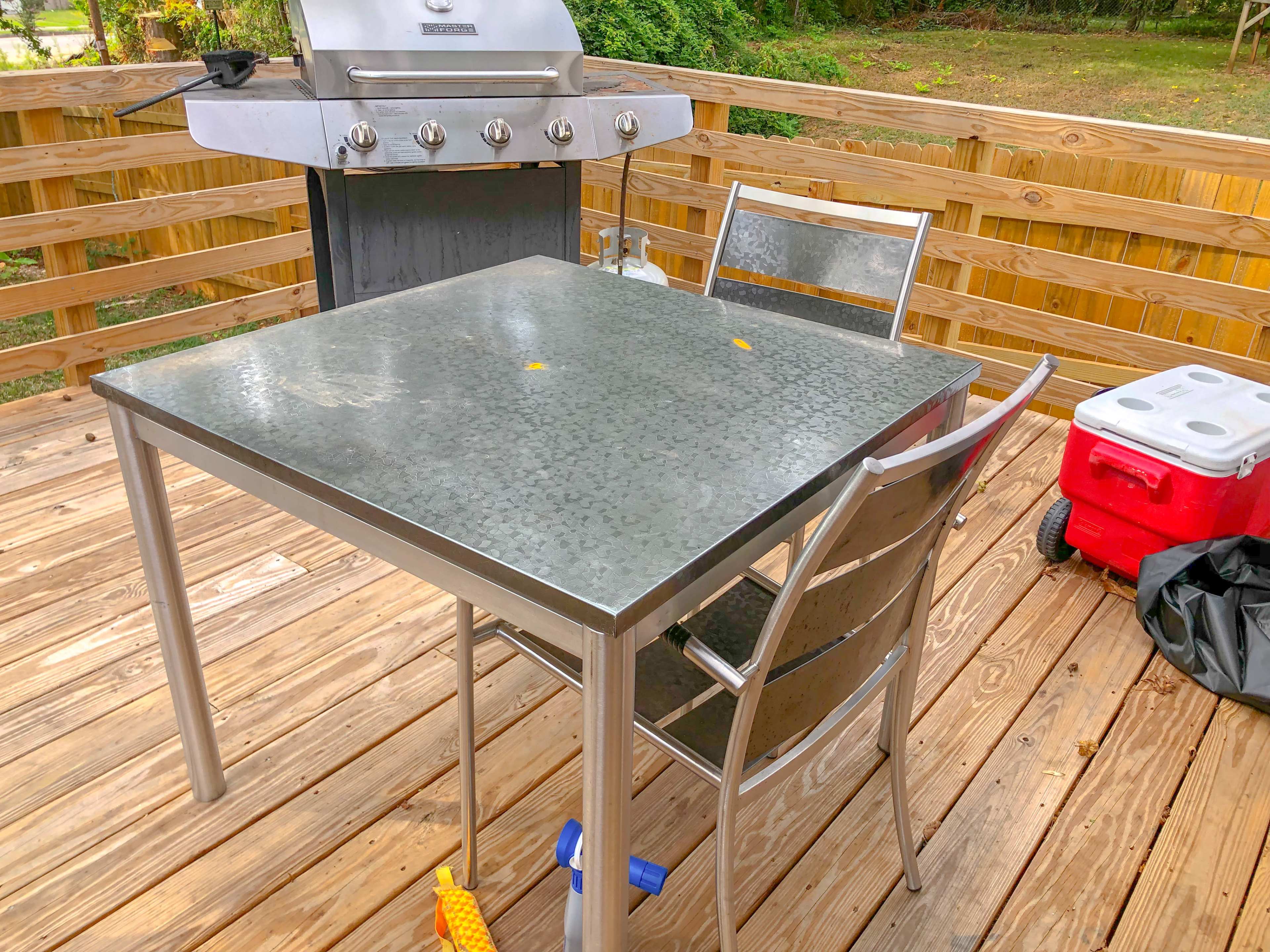 The image shows a stainless steel table and chairs on a wooden deck, with a grill in the background and a cooler to the side.