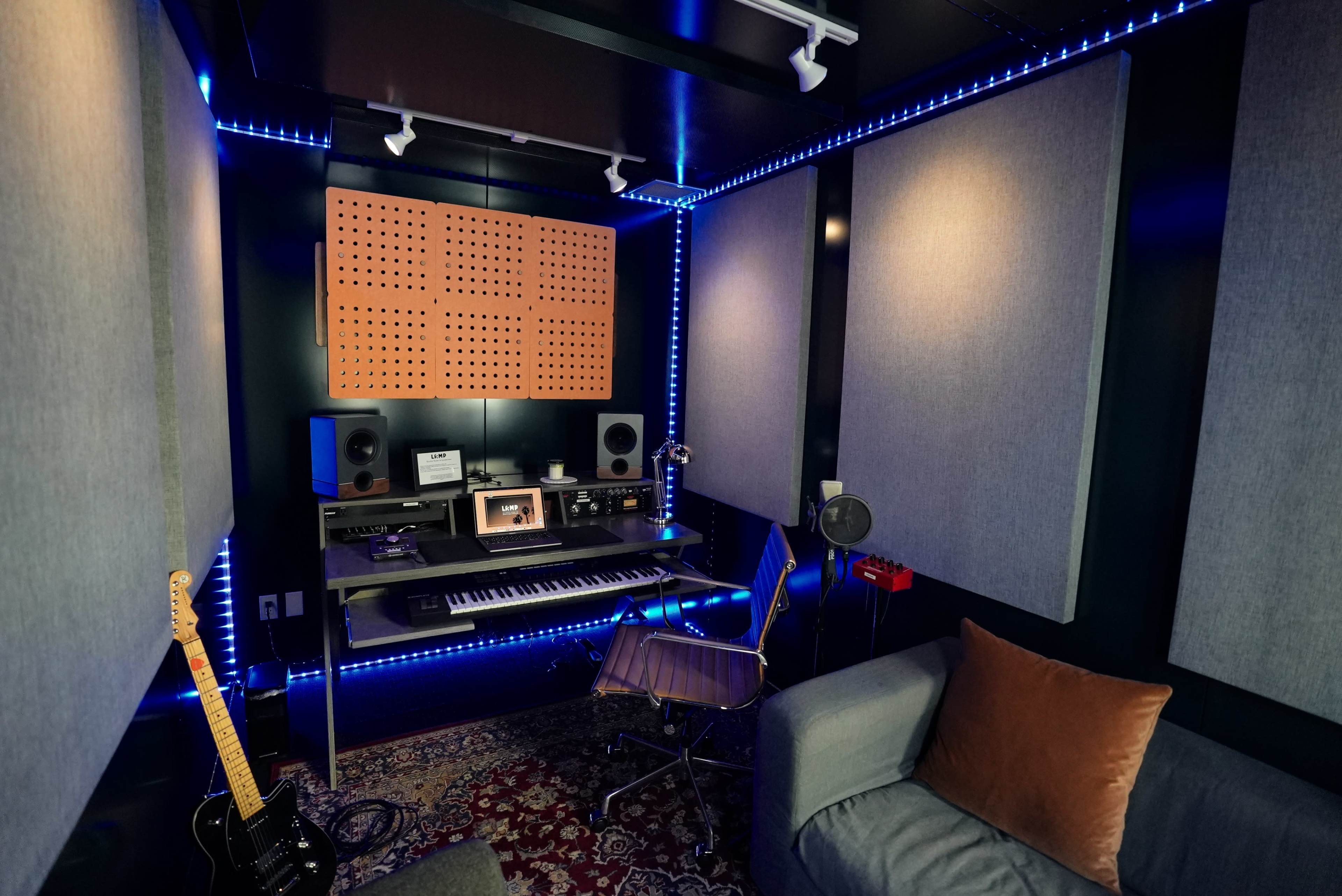 The image shows a compact music studio featuring soundproofing panels, a keyboard, speakers, and a guitar, illuminated by LED lights.