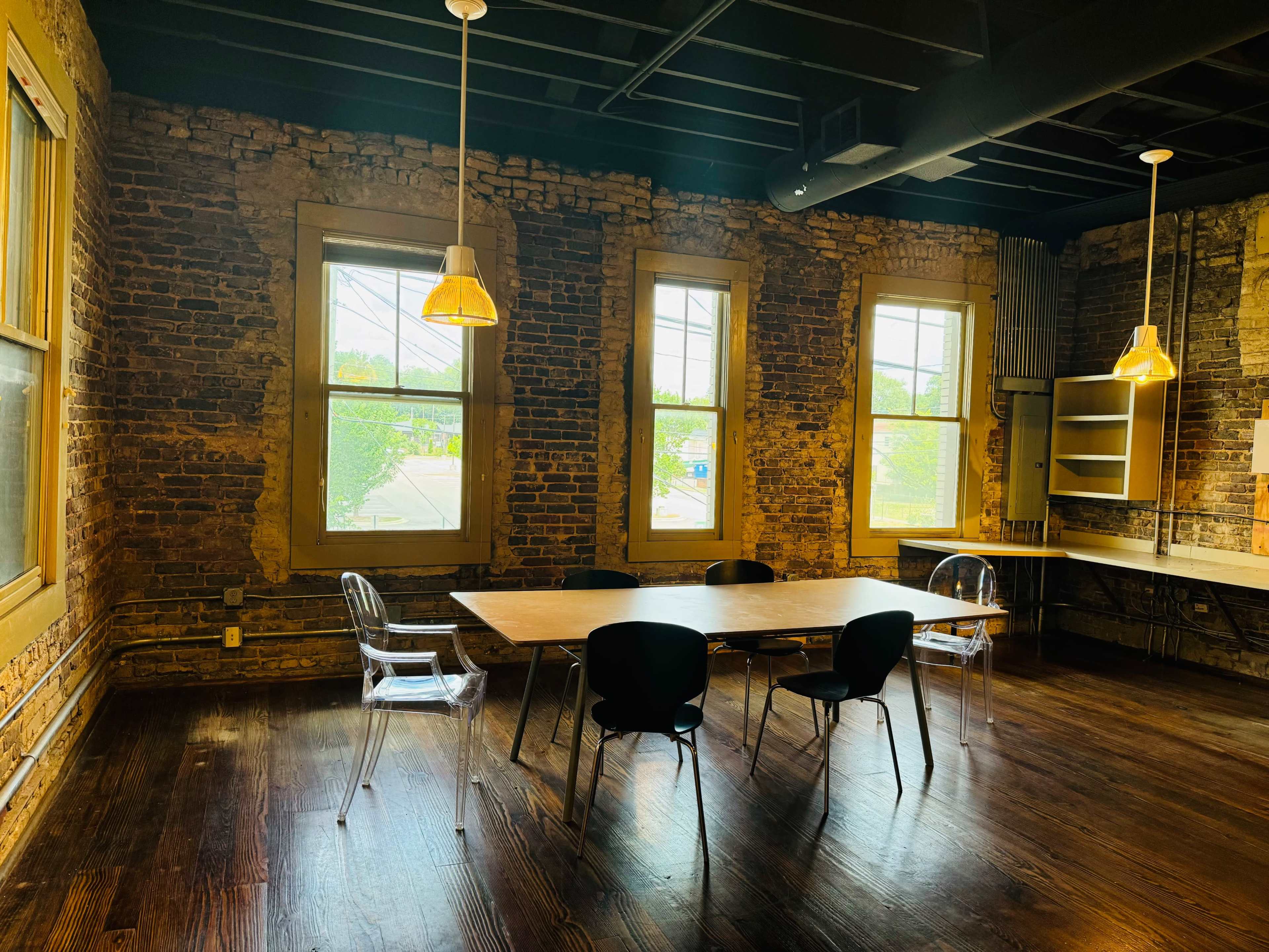 A workspace featuring a wooden table surrounded by black chairs, with exposed brick walls and large windows allowing natural light to enter.