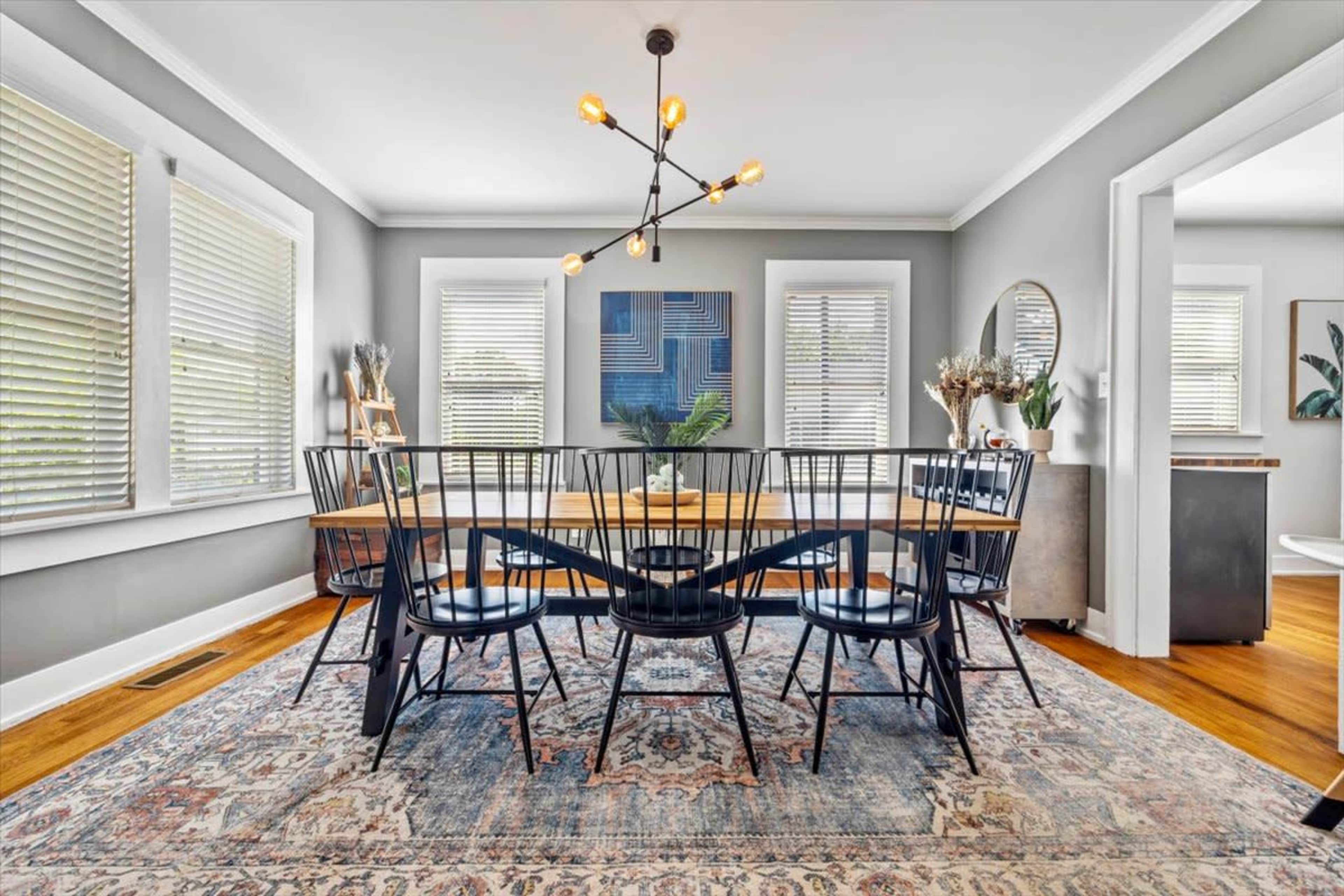 A modern dining room features a large table surrounded by black chairs, with large windows allowing natural light and a patterned rug on the wooden floor.