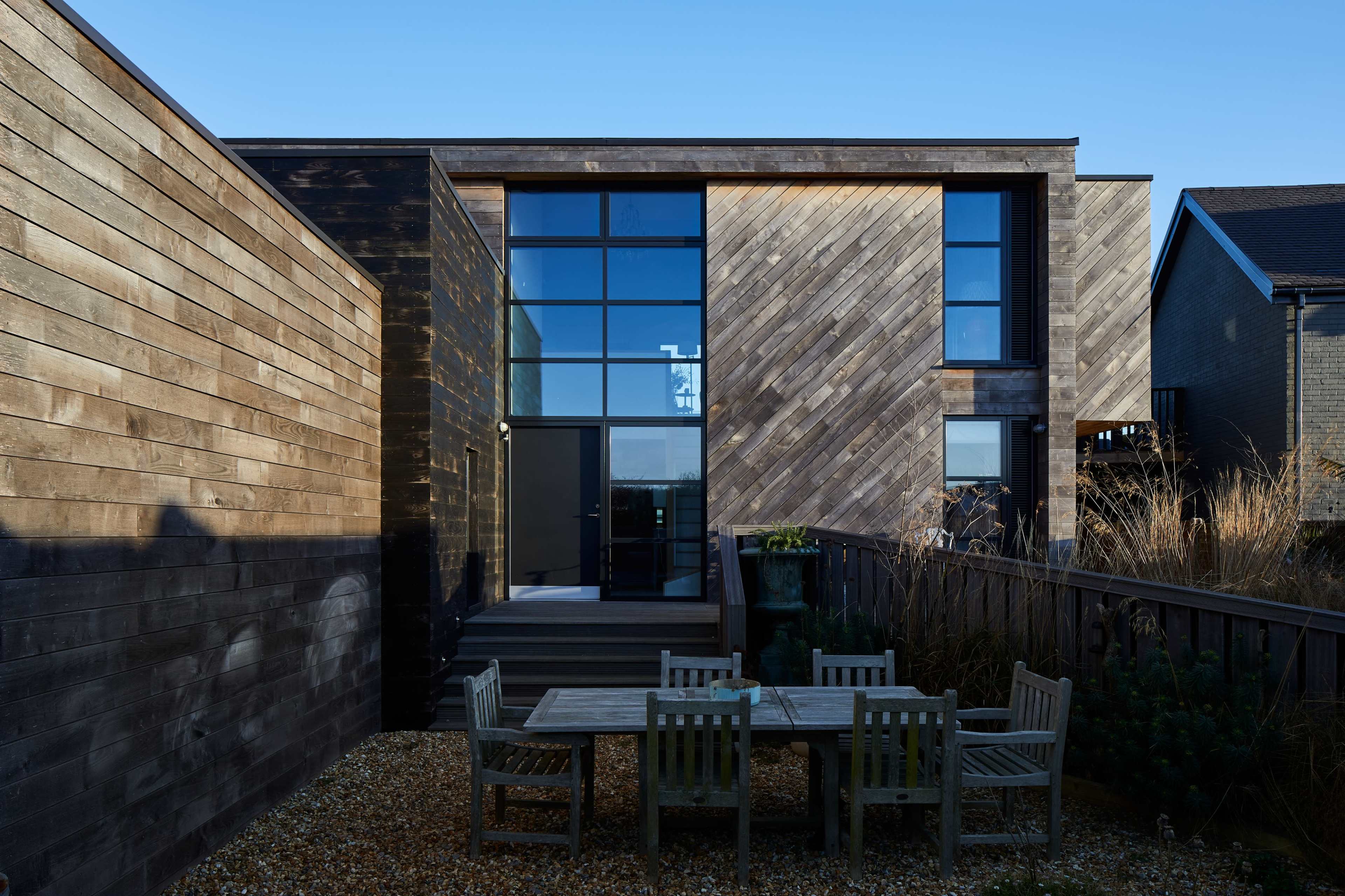 A modern, two-story wooden house with large windows and a patio area featuring a table and chairs, surrounded by gravel and plants.